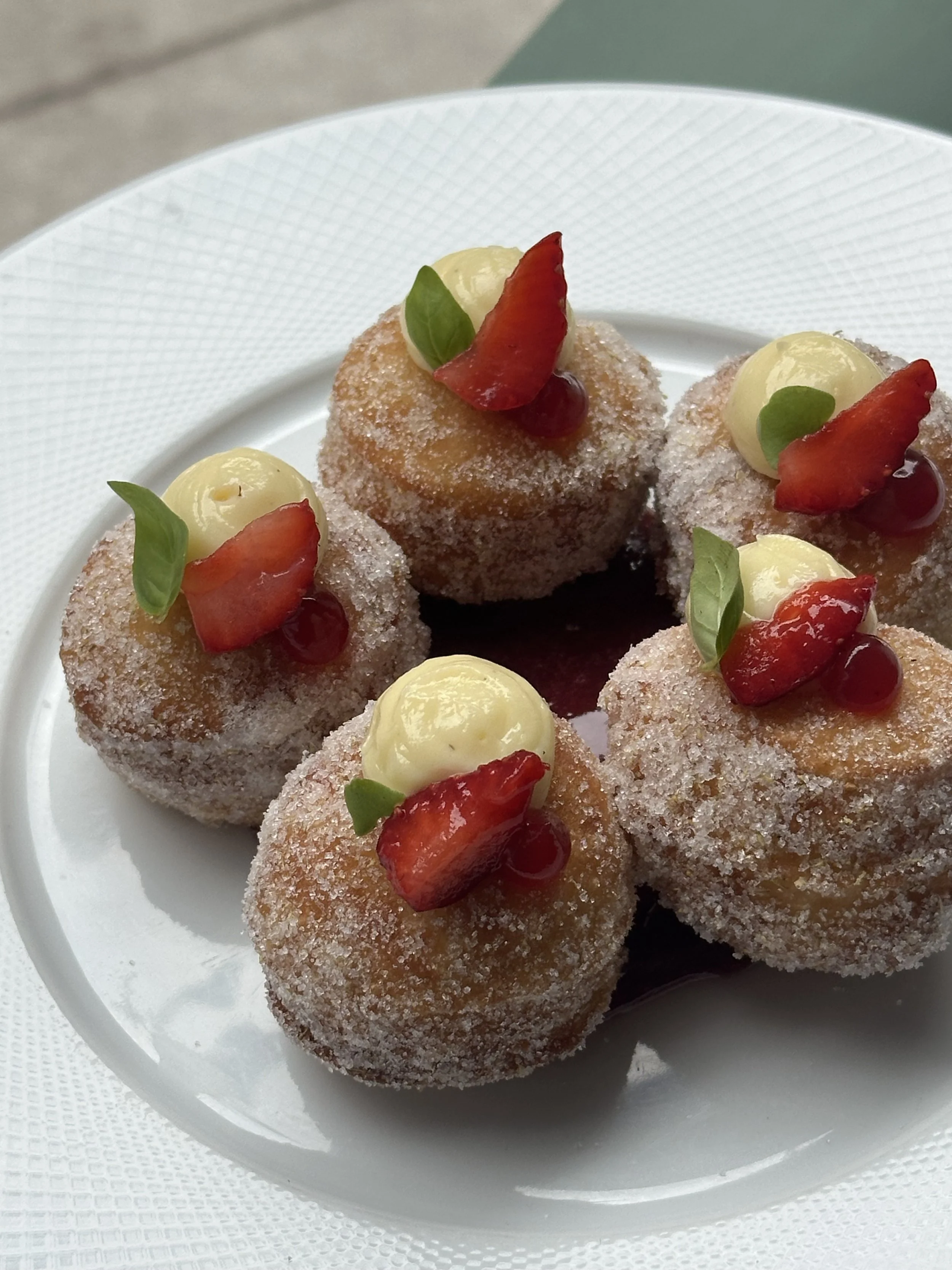Plate of six small, round dessert balls coated with sugar, topped with cream, sliced strawberries, small green leaves, and red berries.