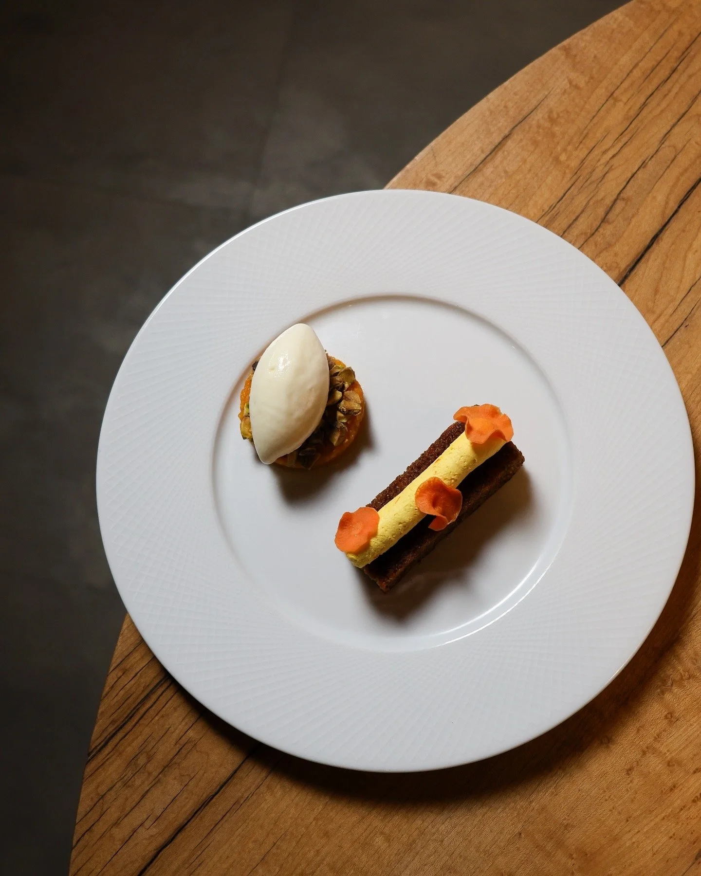 Elegant plated dessert with a scoop of vanilla ice cream on a bed of nuts and caramel, and a rectangular layered cake with orange flower-shaped decorations.