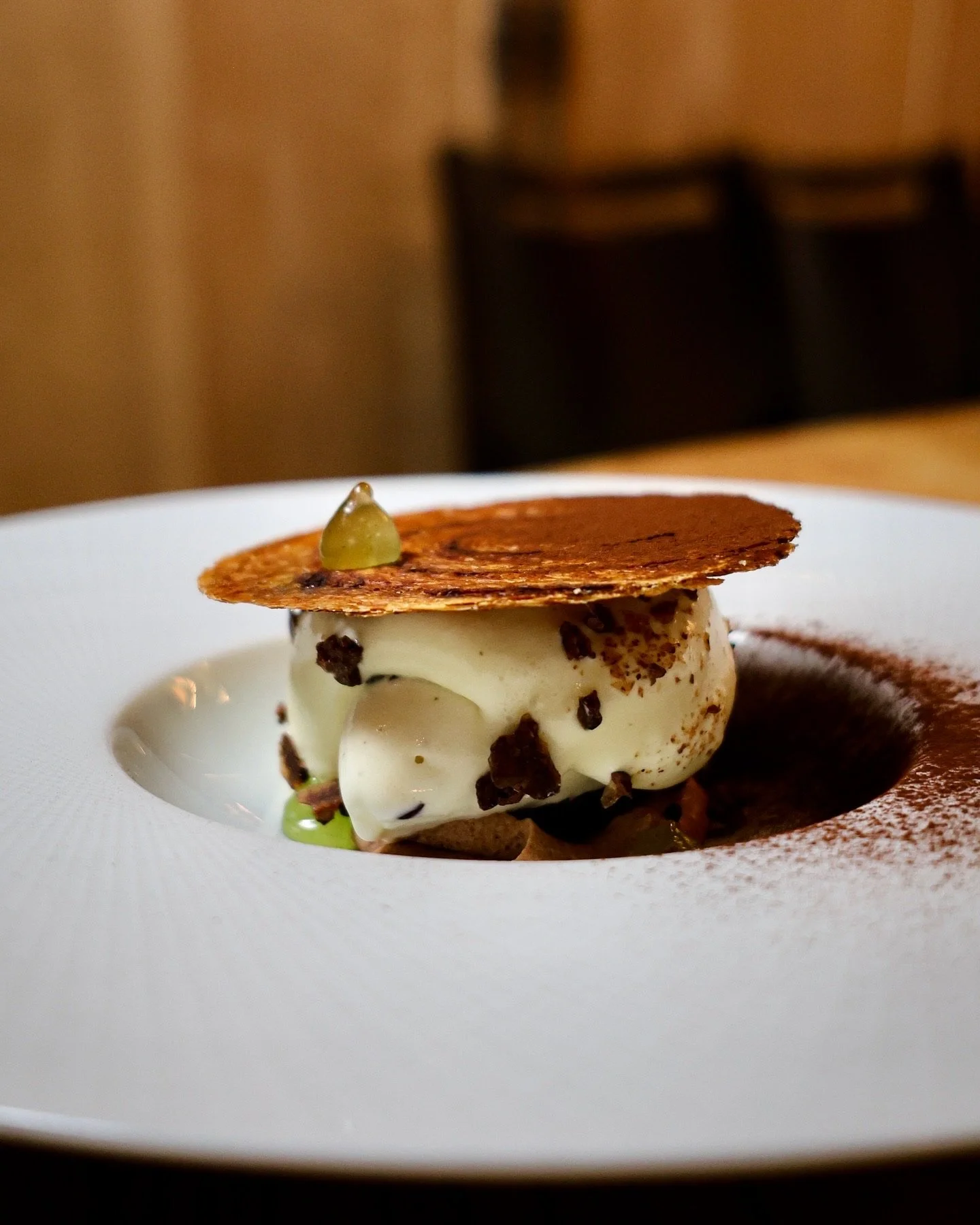 A plated dessert with a scoop of vanilla ice cream, chocolate chunks, a thin crispy cookie on top, and a small green garnish on the cookie.