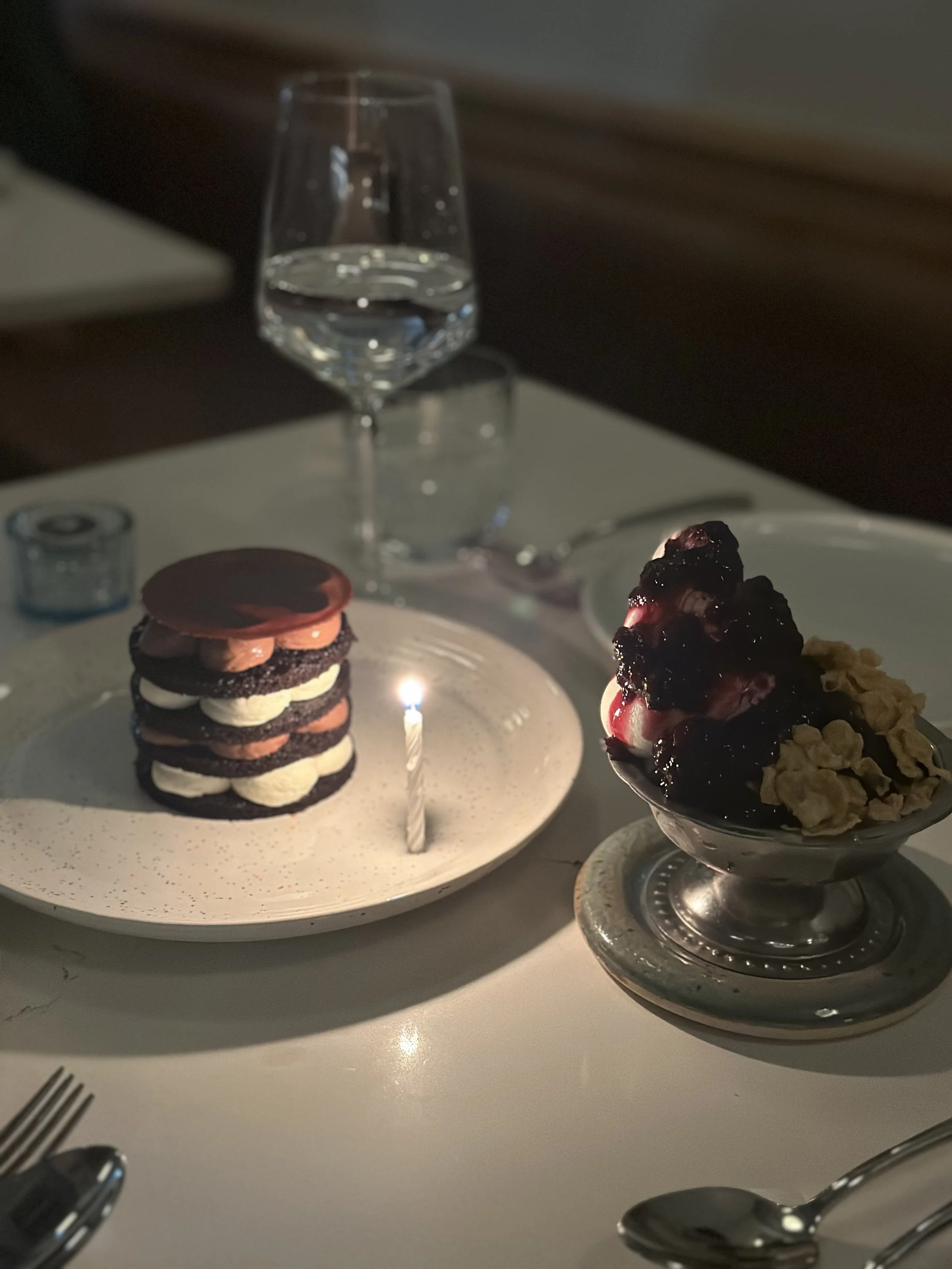 A dinner table with a glass of water, a small dessert with a lit candle, and a bowl of ice cream topped with berries and cereal.