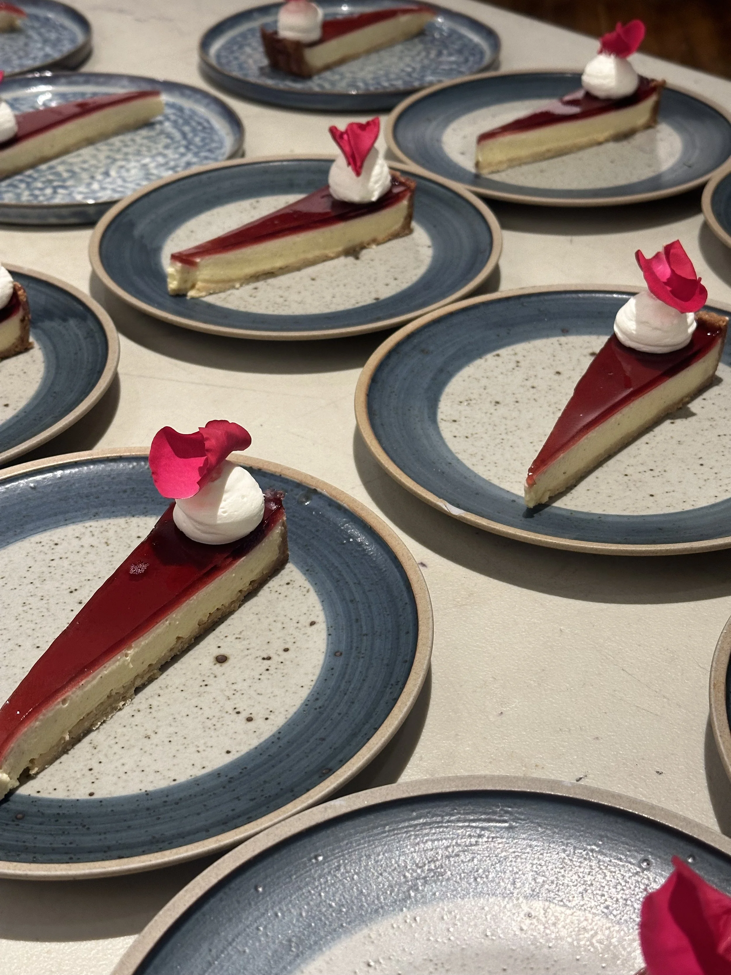 Multiple plates of cheesecake with a red glaze, topped with whipped cream and pink flower petals.