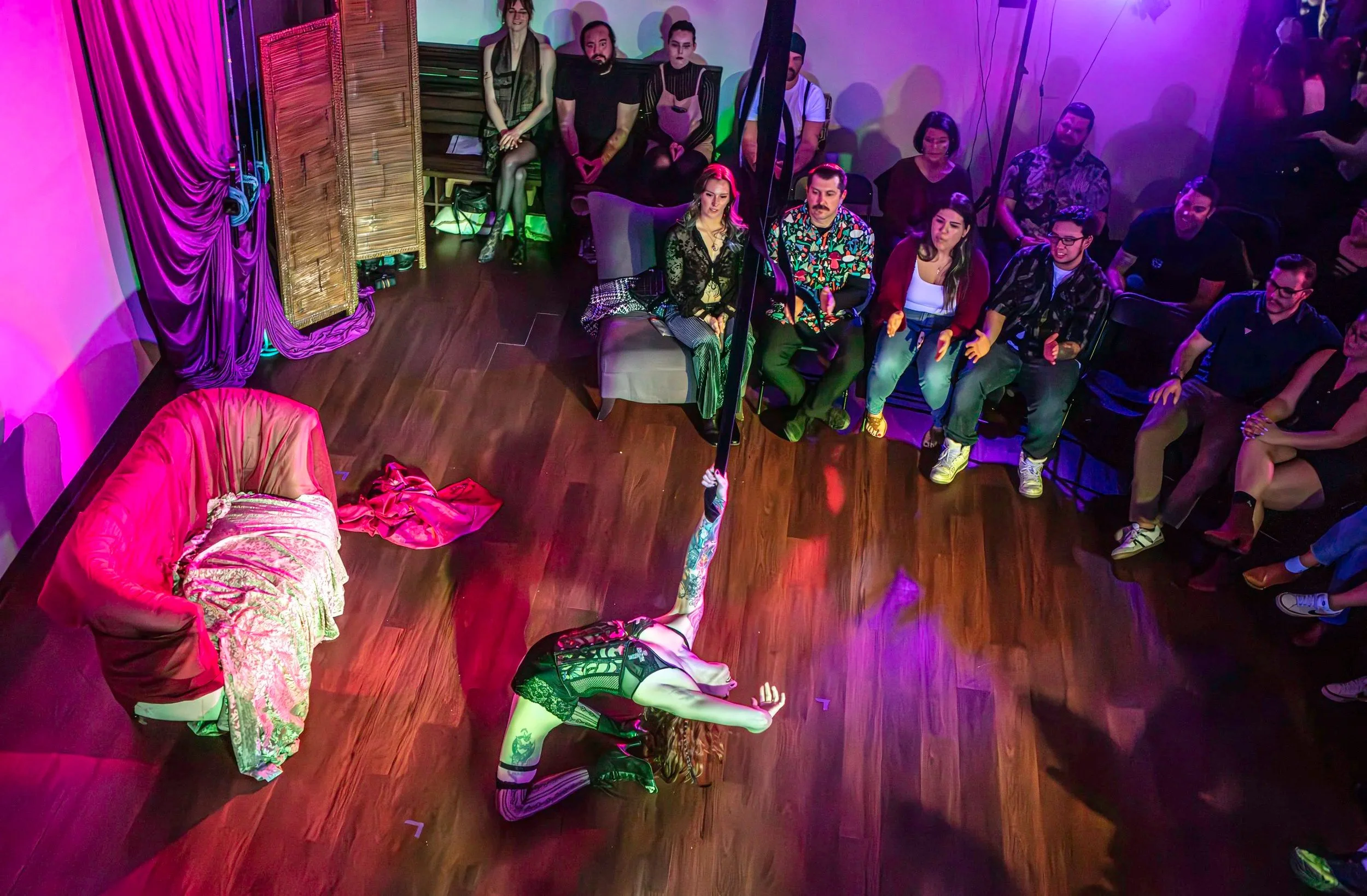 A woman wearing a black lace top and plaid pants performs a dance move on the floor in a room with purple and pink lighting. Audience members seated and standing watch her performance.