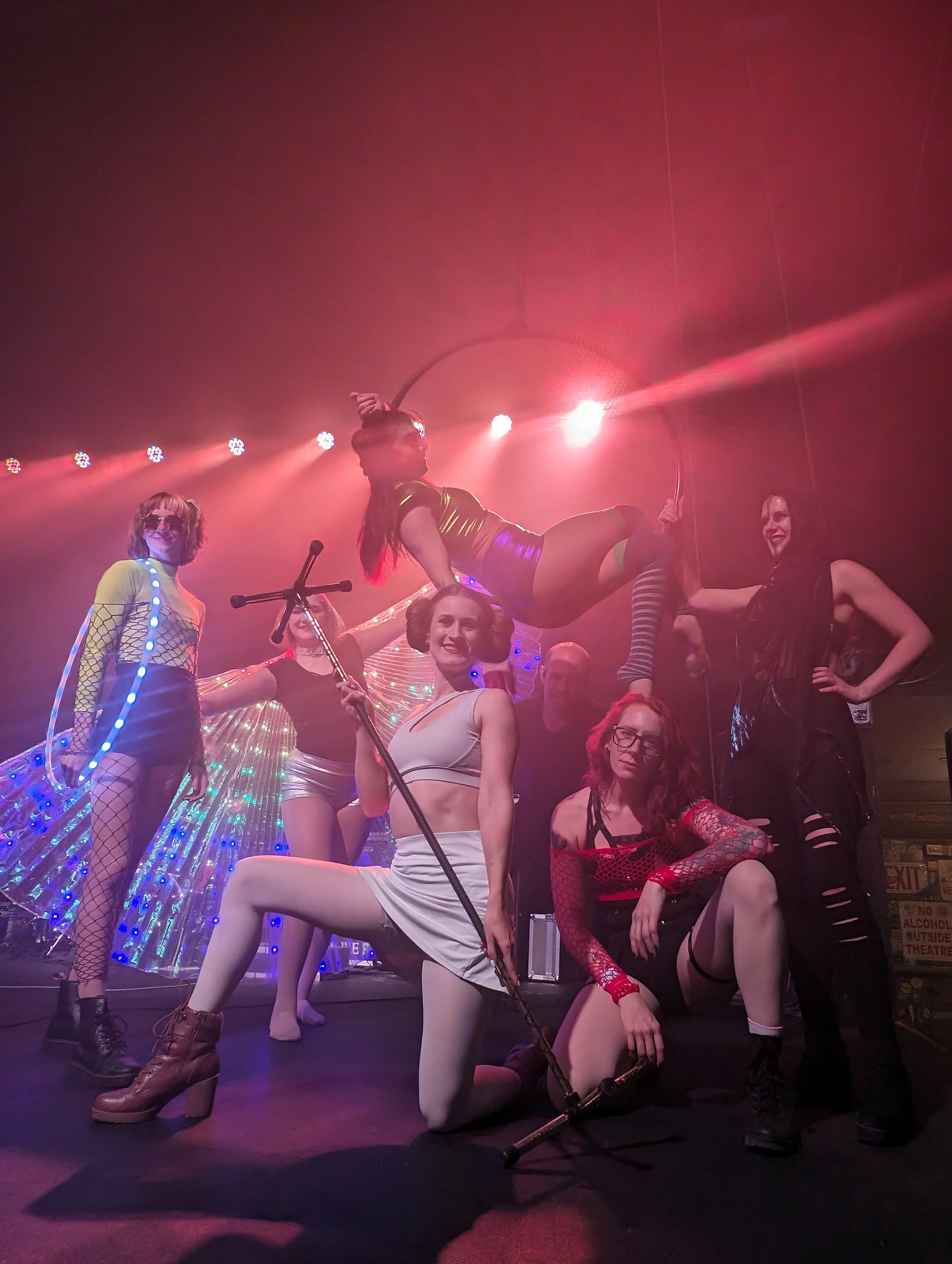 Group of performers on stage in colorful costumes with neon lights, some sitting and others standing or posing in energetic positions.