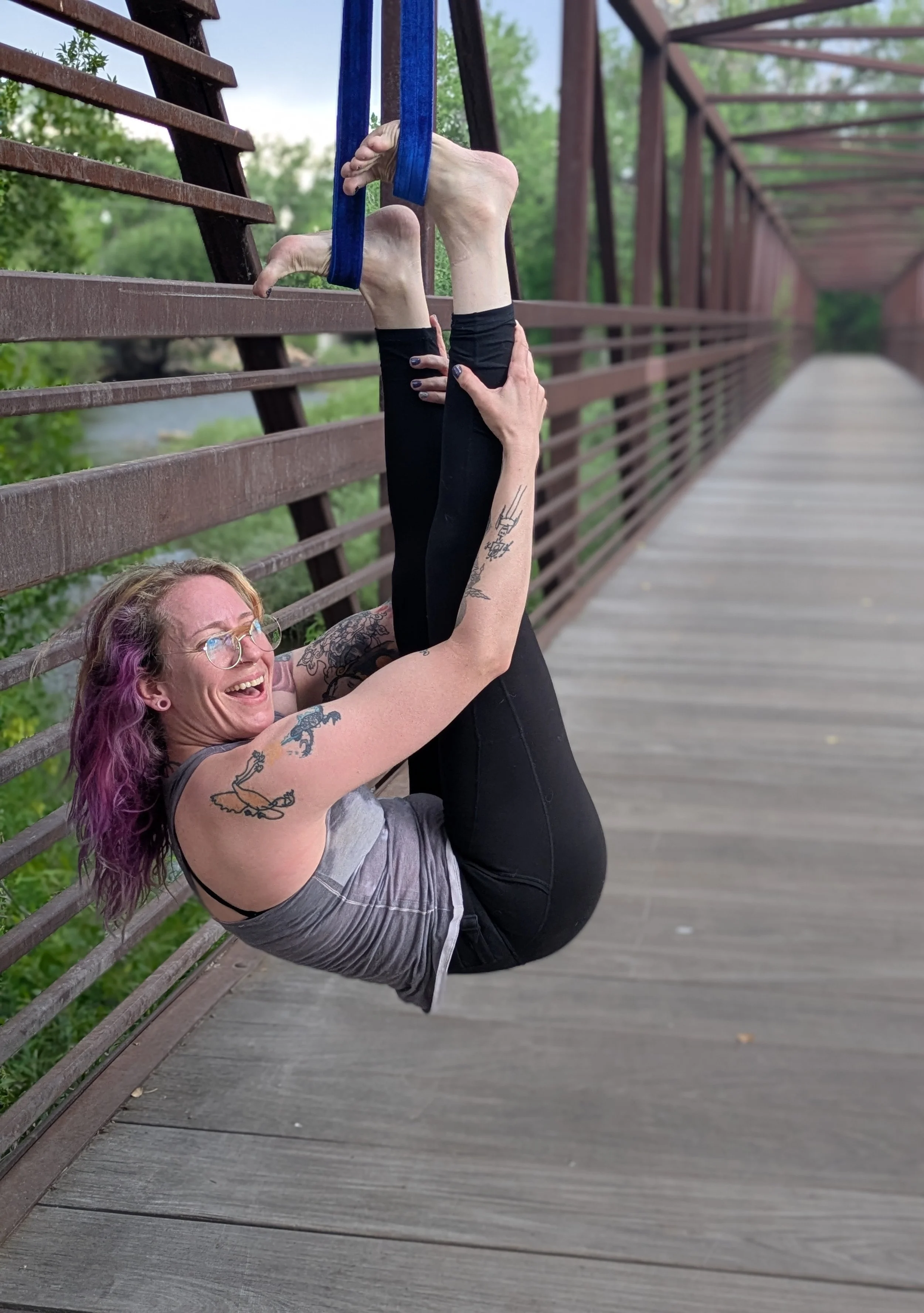 A woman with tattoos and glasses practicing yoga on a bridge, holding her legs in the air with a yoga strap, smiling