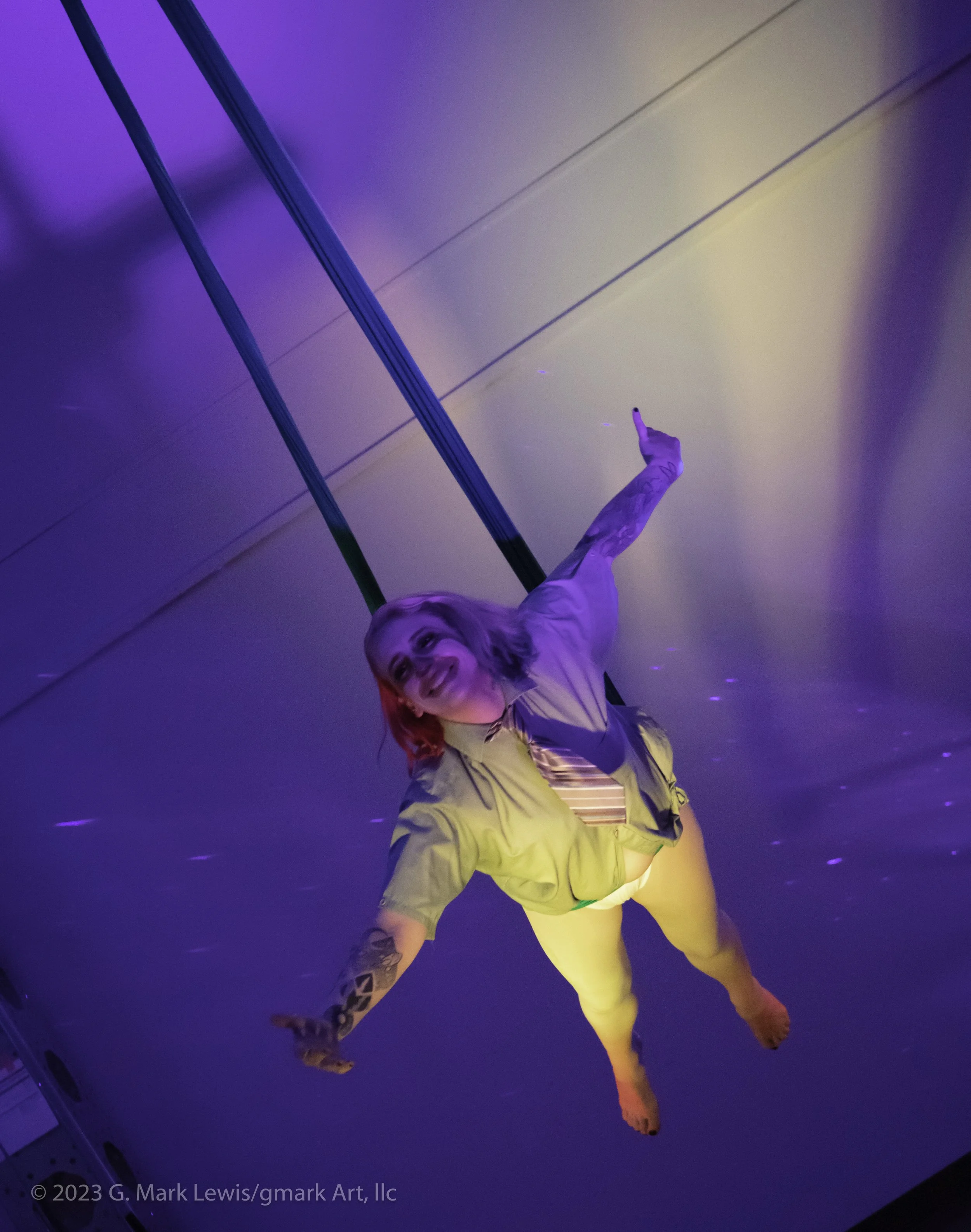 A woman with red hair in a yellow outfit is dancing or performing, with her arm outstretched and her head tilted back, in a room illuminated with purple and yellow lighting.