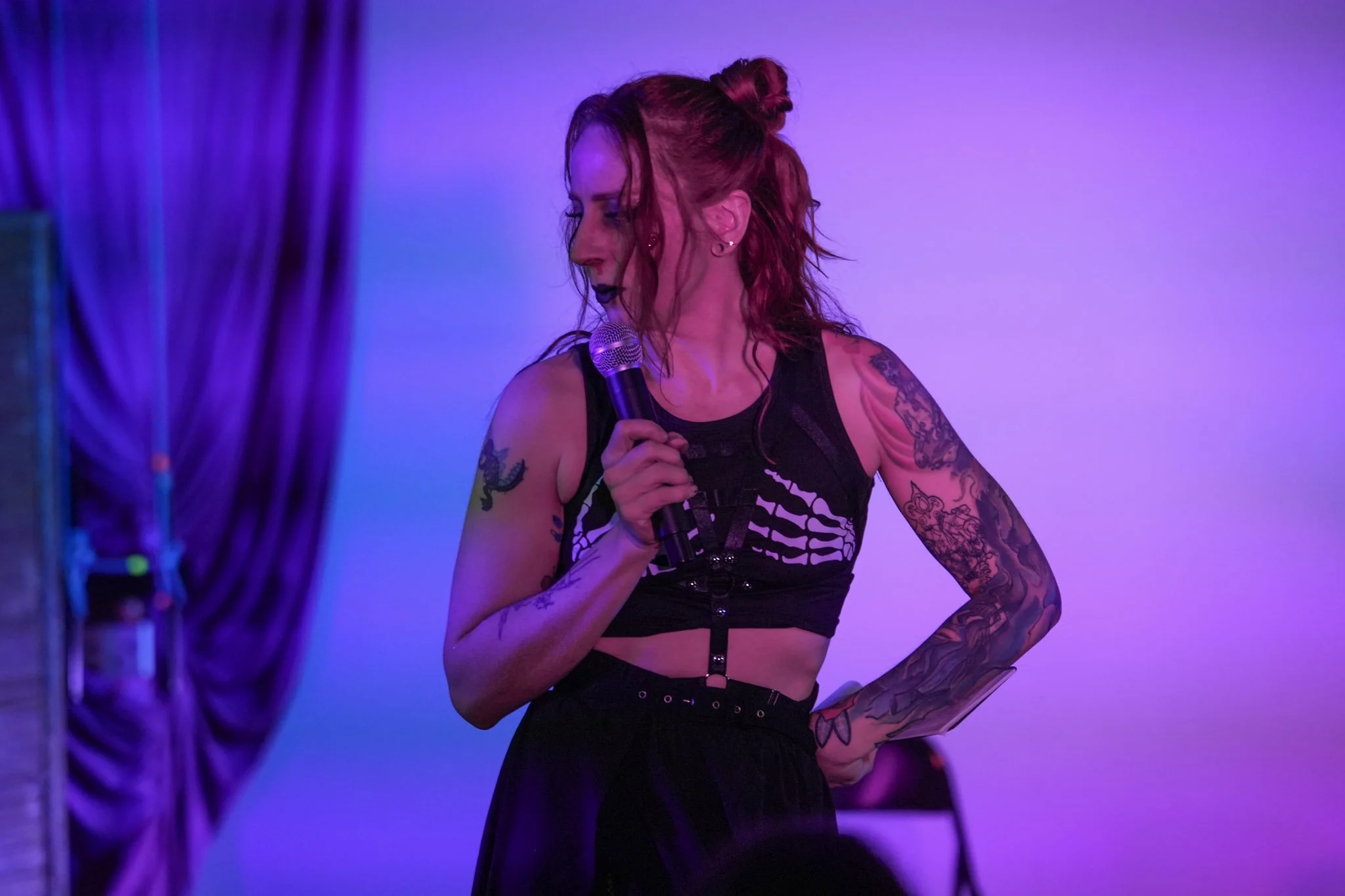 A woman with red hair, tattoos on her arms, wearing a black crop top, holding a microphone, on stage with purple lighting.