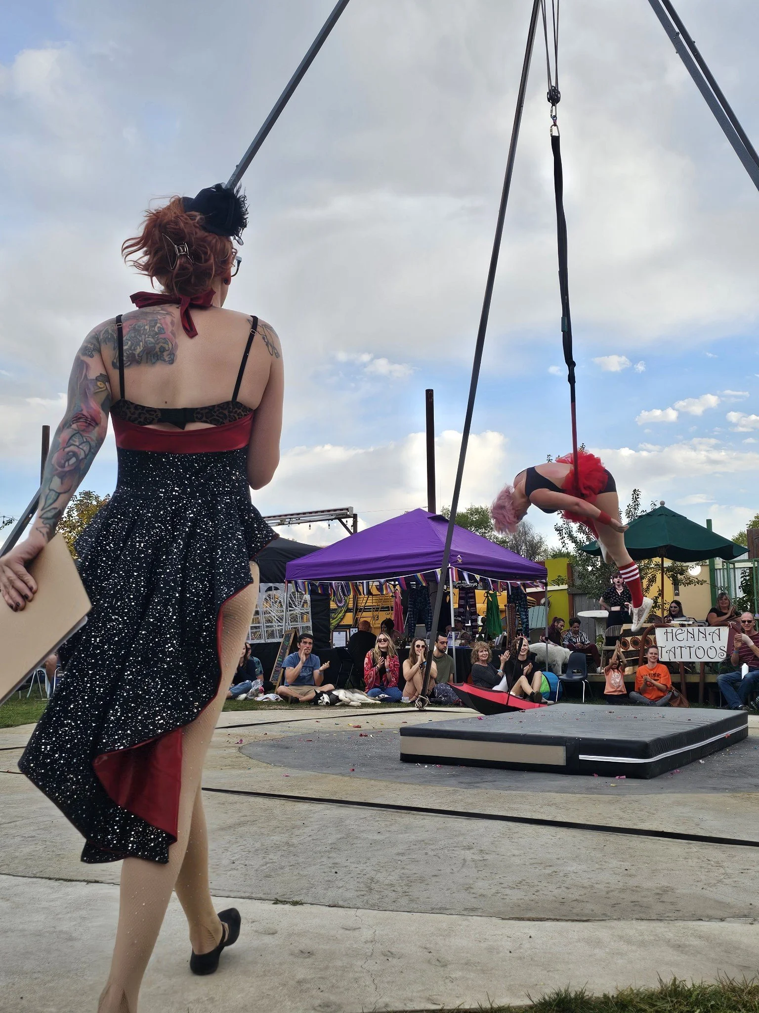 A performer with pink hair and a red tutu suspends upside down from a trapeze at an outdoor event with an audience seated on the ground.