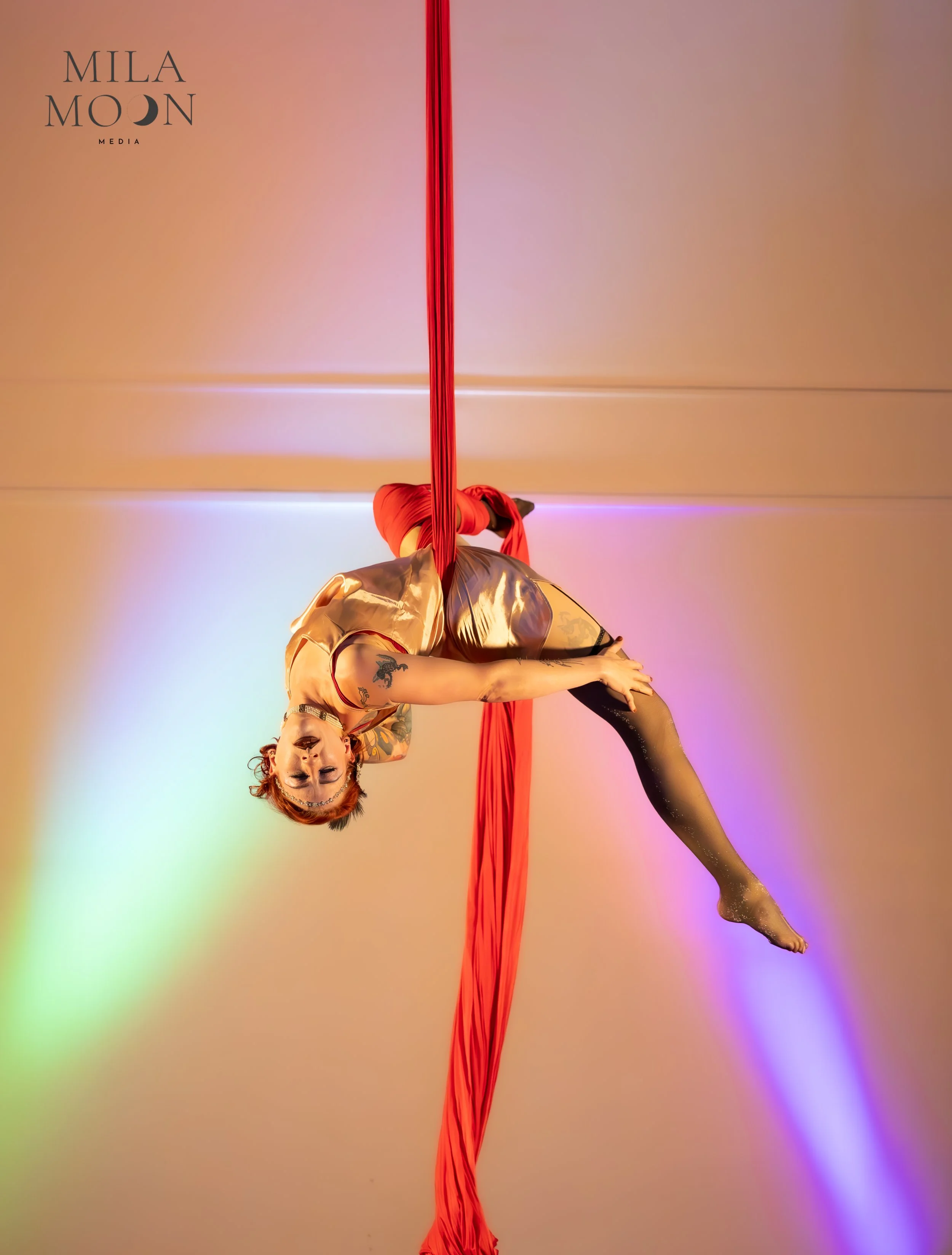 A person with short red hair and tattoos is performing an aerial silk routine, hanging upside down with one leg extended and arms touching the floor, against a pastel, gradient background.