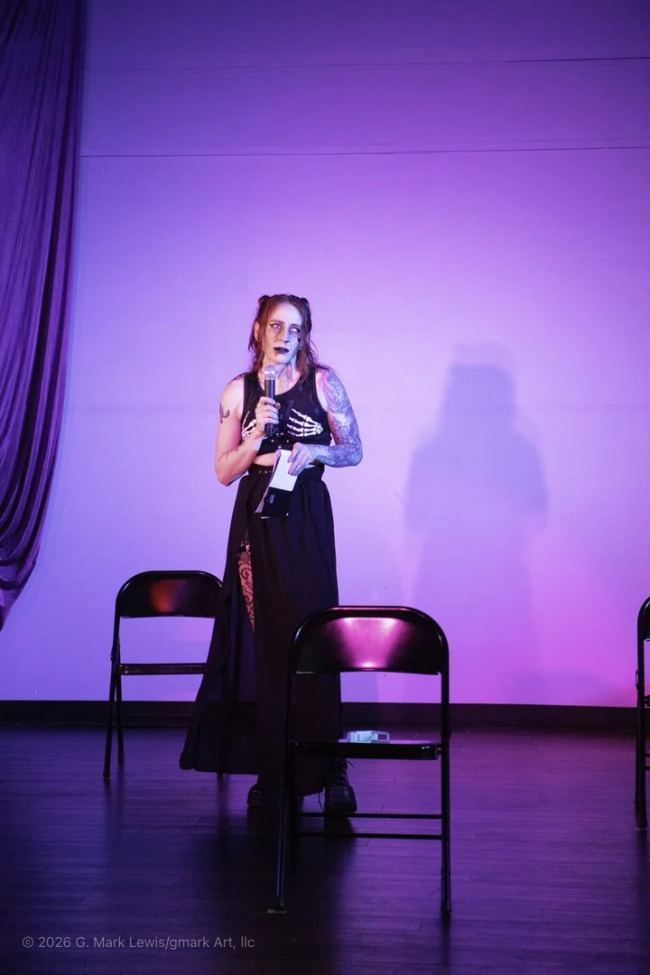 A young woman with tattoos and dark makeup holding a microphone and a piece of paper, standing on a stage with purple and pink lighting, behind three empty chairs.