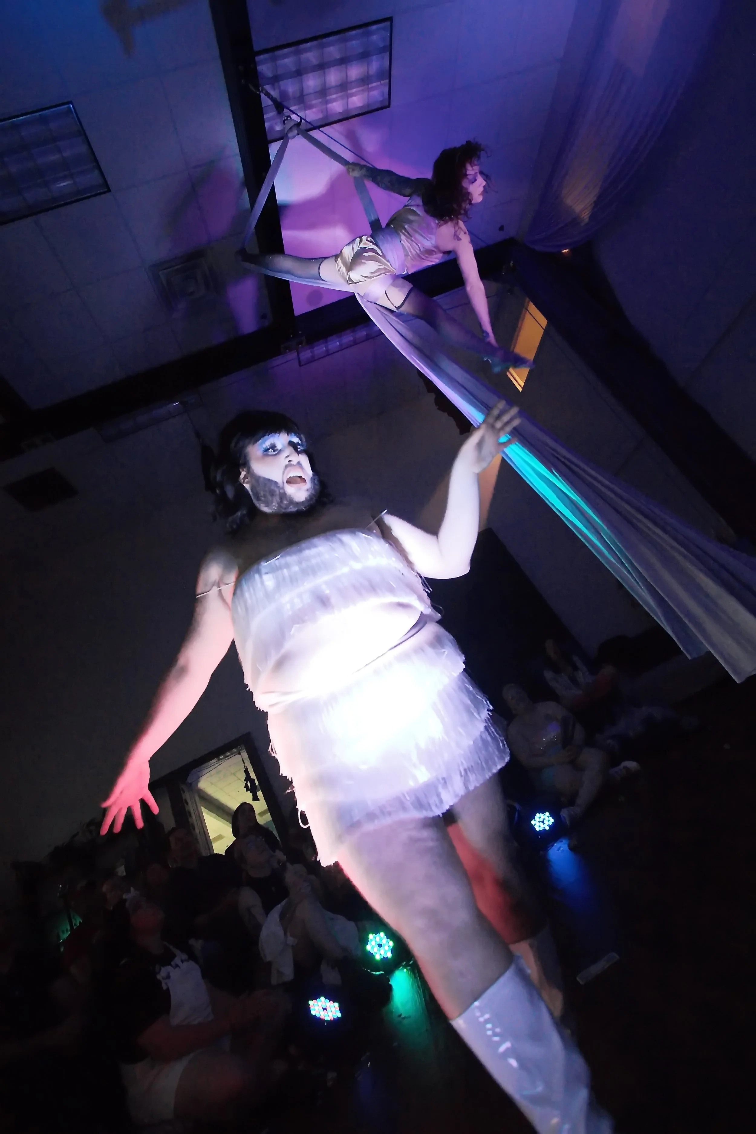 Performer in a shiny, light-colored dress and knee-high boots on stage, with an audience behind, under purple and green stage lights.