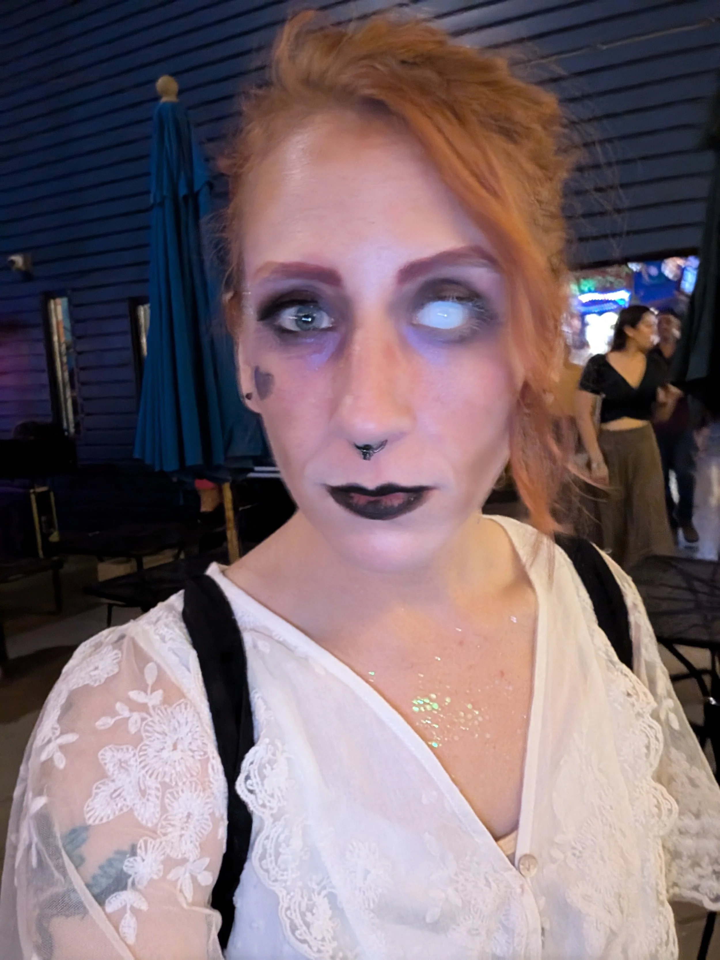 A woman with red hair and makeup resembling a skeleton face, wearing a white lace top, at a social gathering or event.