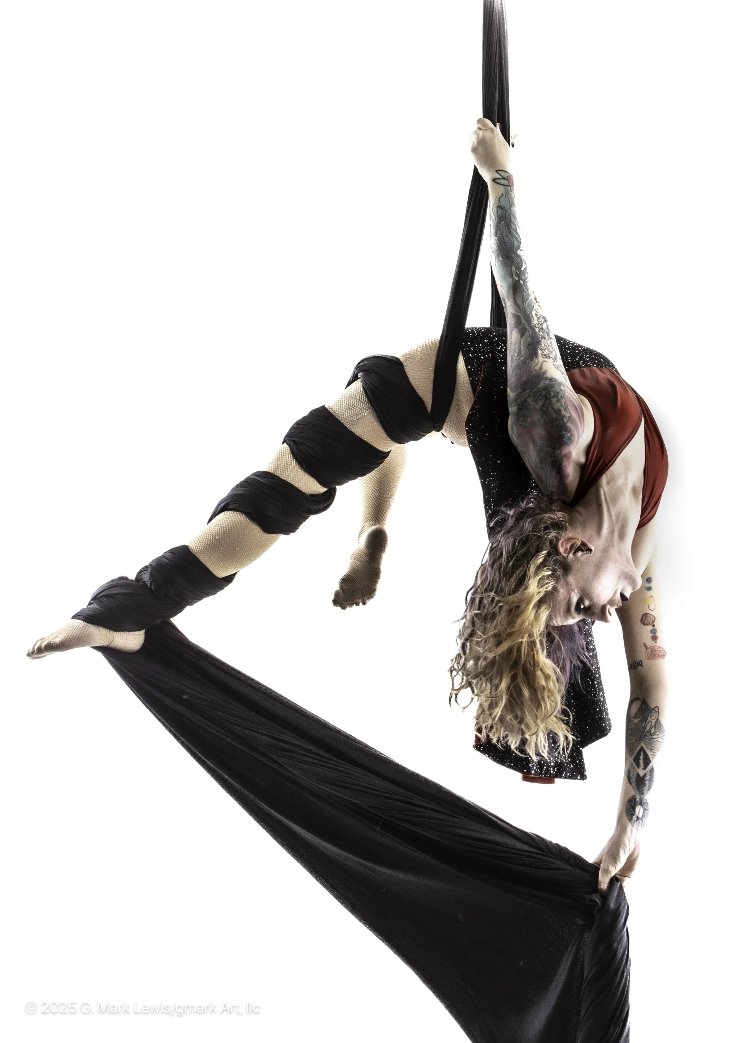 A woman performing aerial silk acrobatics, hanging upside down with one leg wrapped around black fabric, wearing a red top, black skirt with white specks, and fishnet stockings. She has tattoos on her arms and long curly hair.