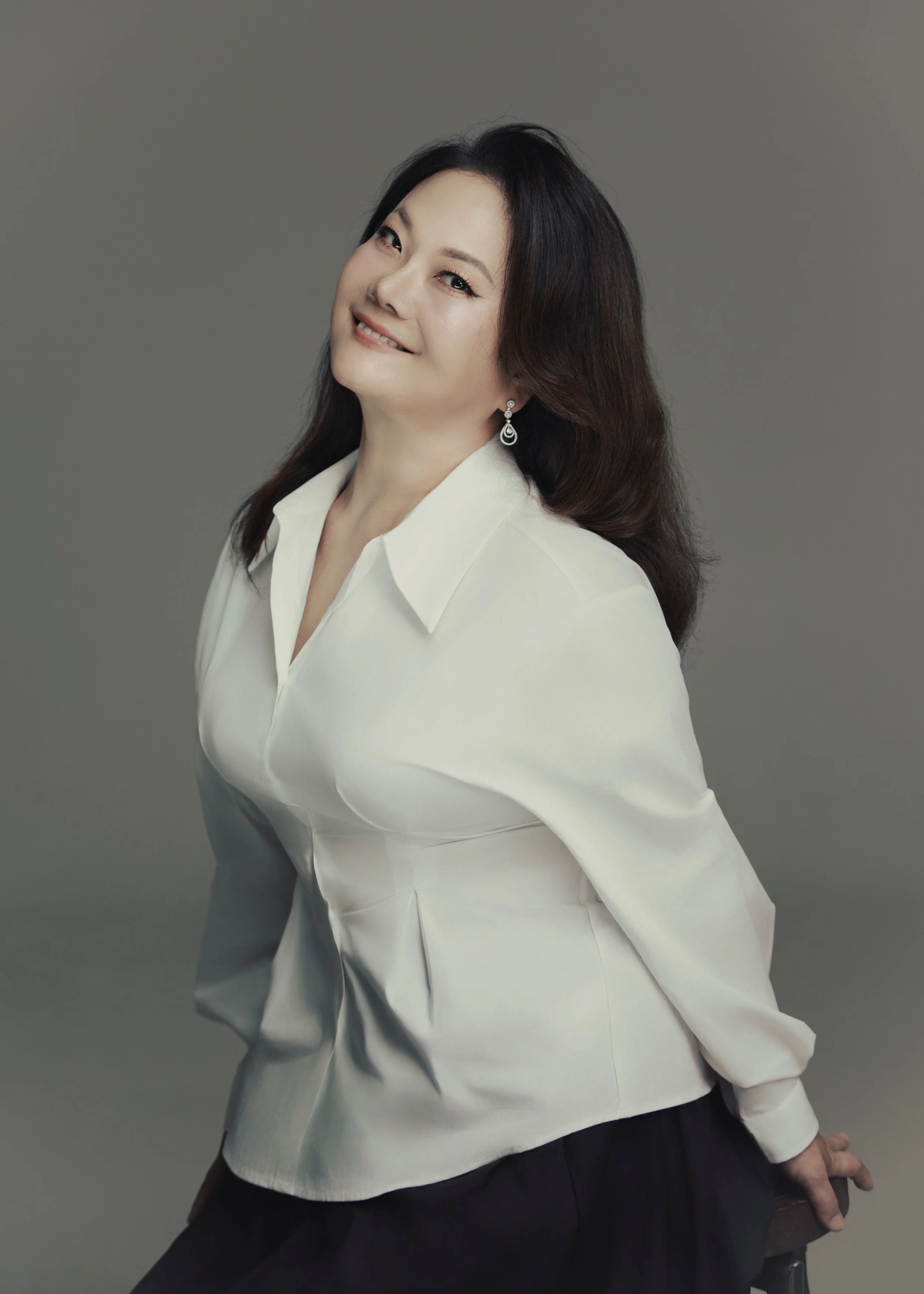 A woman with long dark brown hair, wearing a white blazer and black pants, posing against a gray background.