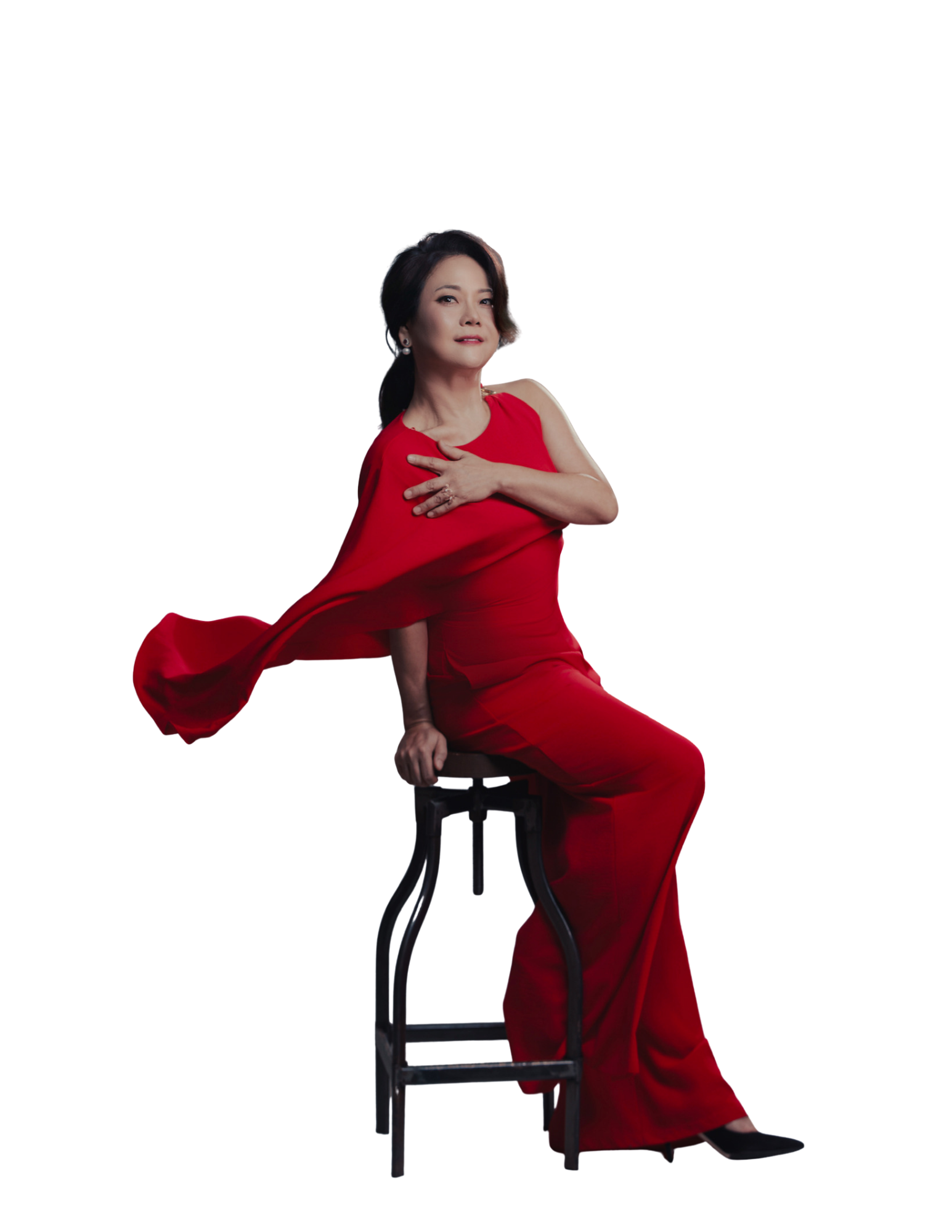 A woman with short dark hair wearing a red dress sits on a stool, with one hand on her shoulder and the other on her lap, against a black background.
