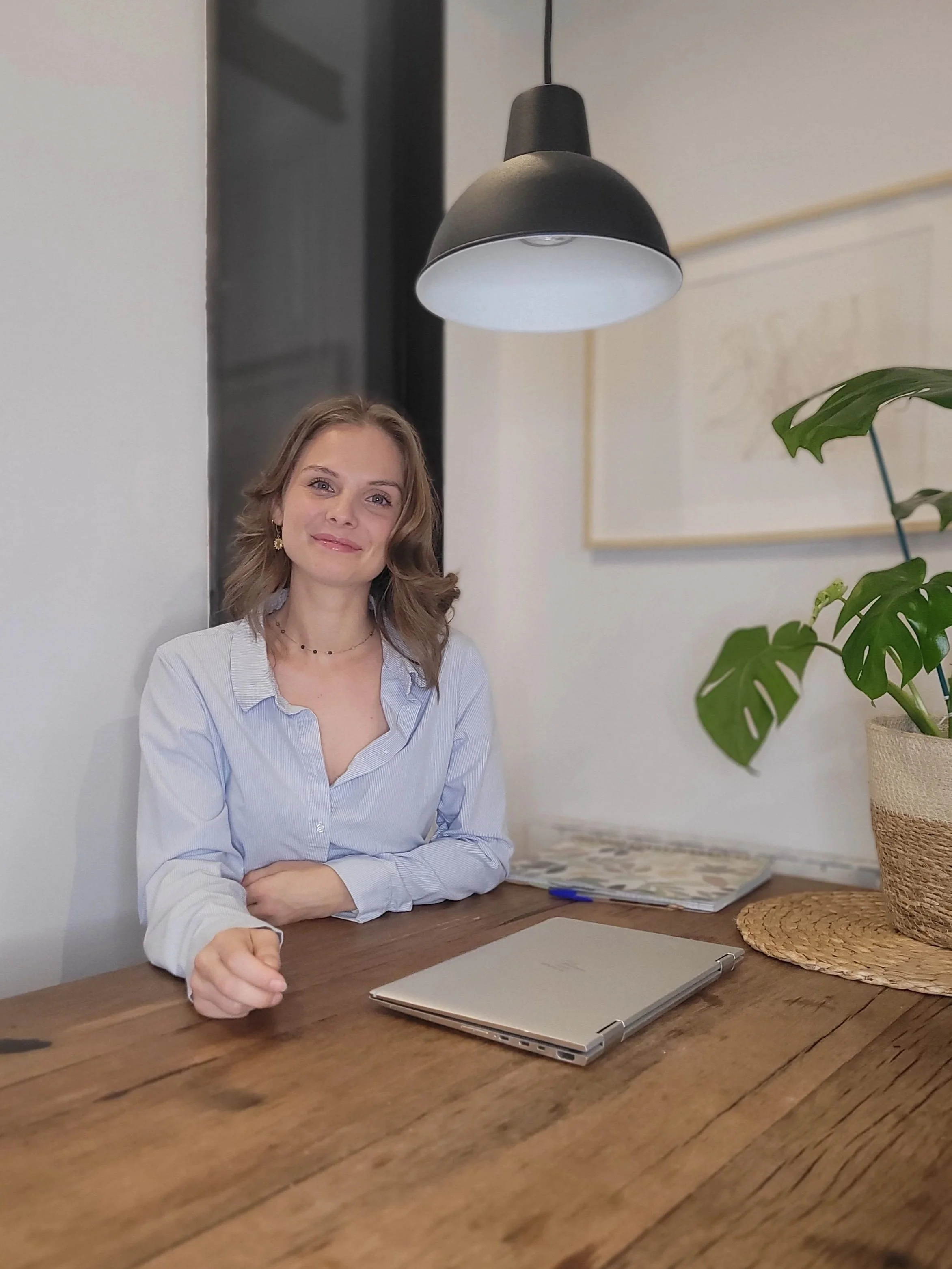 Une femme souriante, assise à une table en bois à l'intérieur, sous un luminaire noir, avec un ordinateur portable fermé, un stylo, et une plante verte