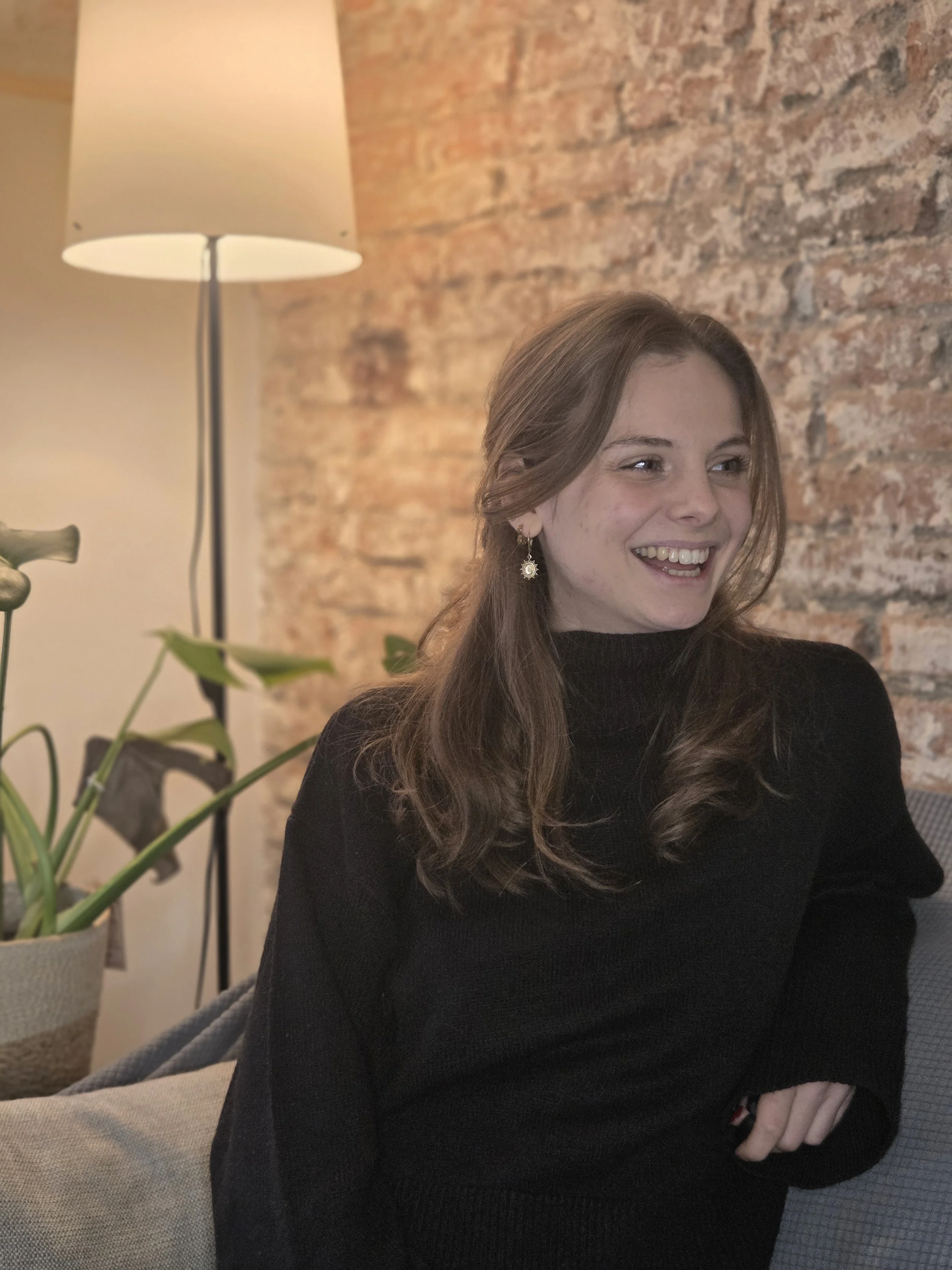 Une jeune femme souriante avec des cheveux bruns, portant un pull noir, assise dans un intérieur avec un mur en brique et une lampe sur pied en arrière-plan.