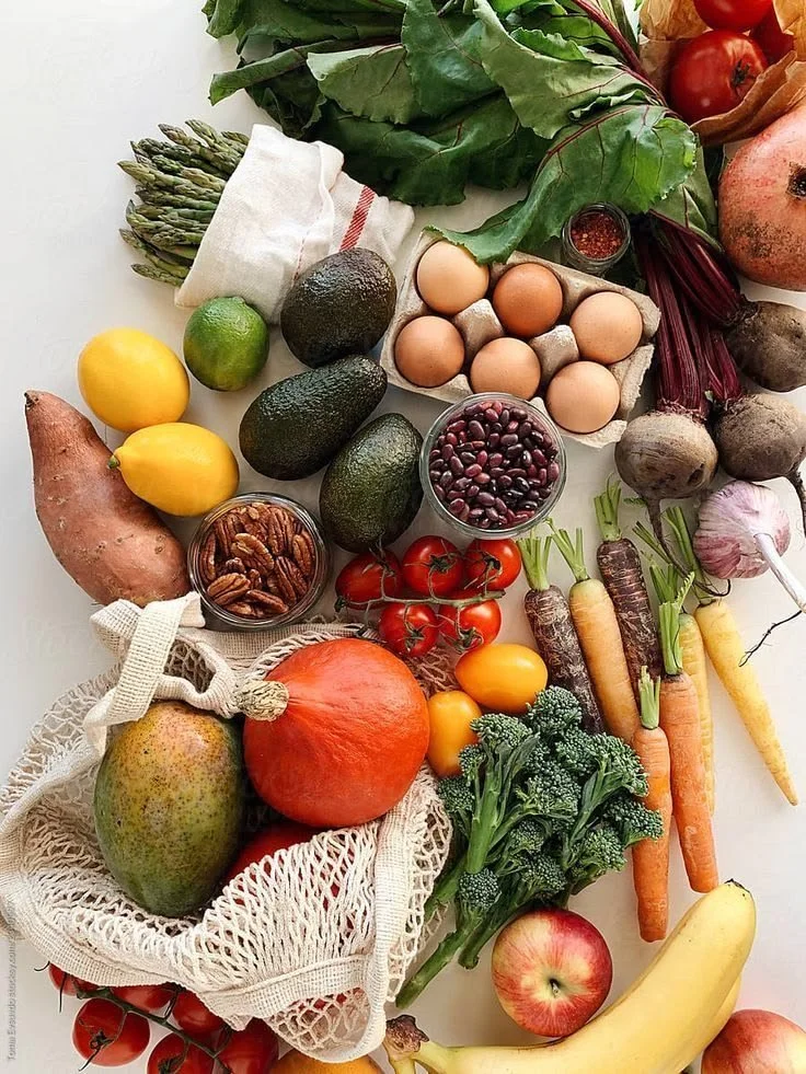 Une variété de fruits et légumes frais, notamment des asperges, avocats, pommes de terre, œufs, tomates, pêches, bananes, carottes, brocoli, betteraves, ail, piment, radis, et une poignée de noix, disposés sur une surface blanche.