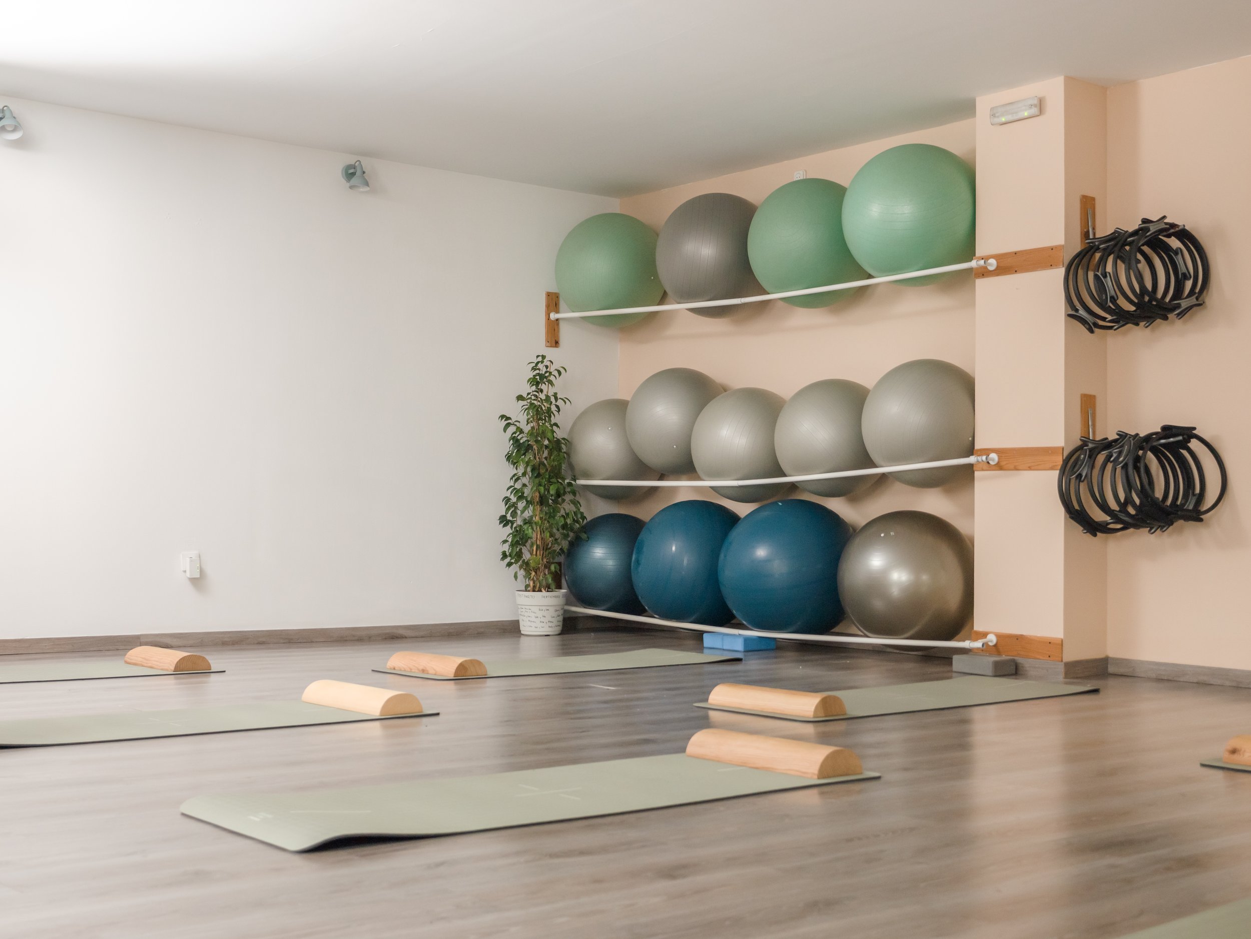 Sala de yoga con colchonetas y rodillos, estantes con pelotas de ejercicio de varios colores, y bandas de resistencia colgadas en la pared a la derecha.