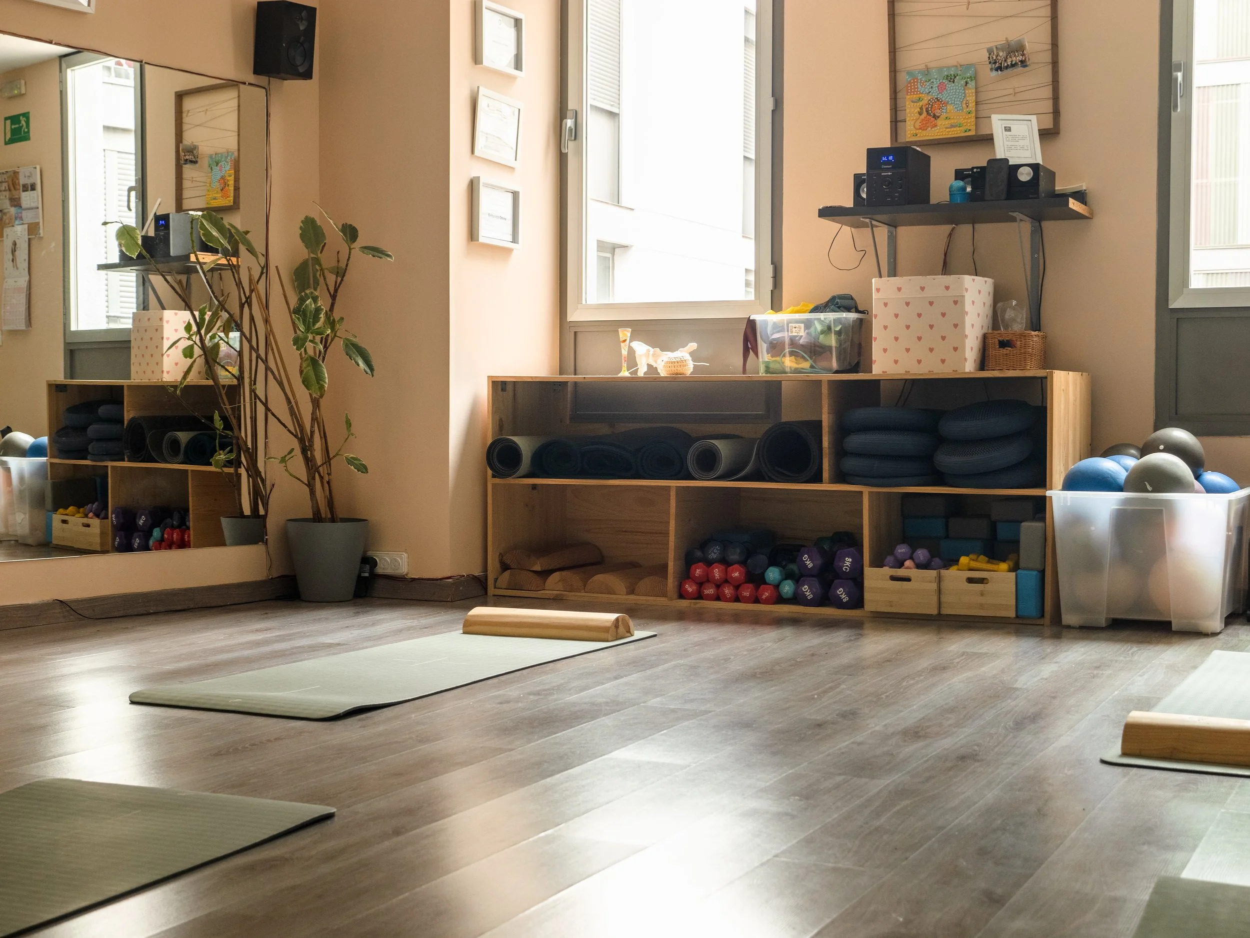 Sala de yoga con colchonetas, pesas y bolas de ejercicio, con una planta en una maceta y ventanas que dejan entrar luz natural.