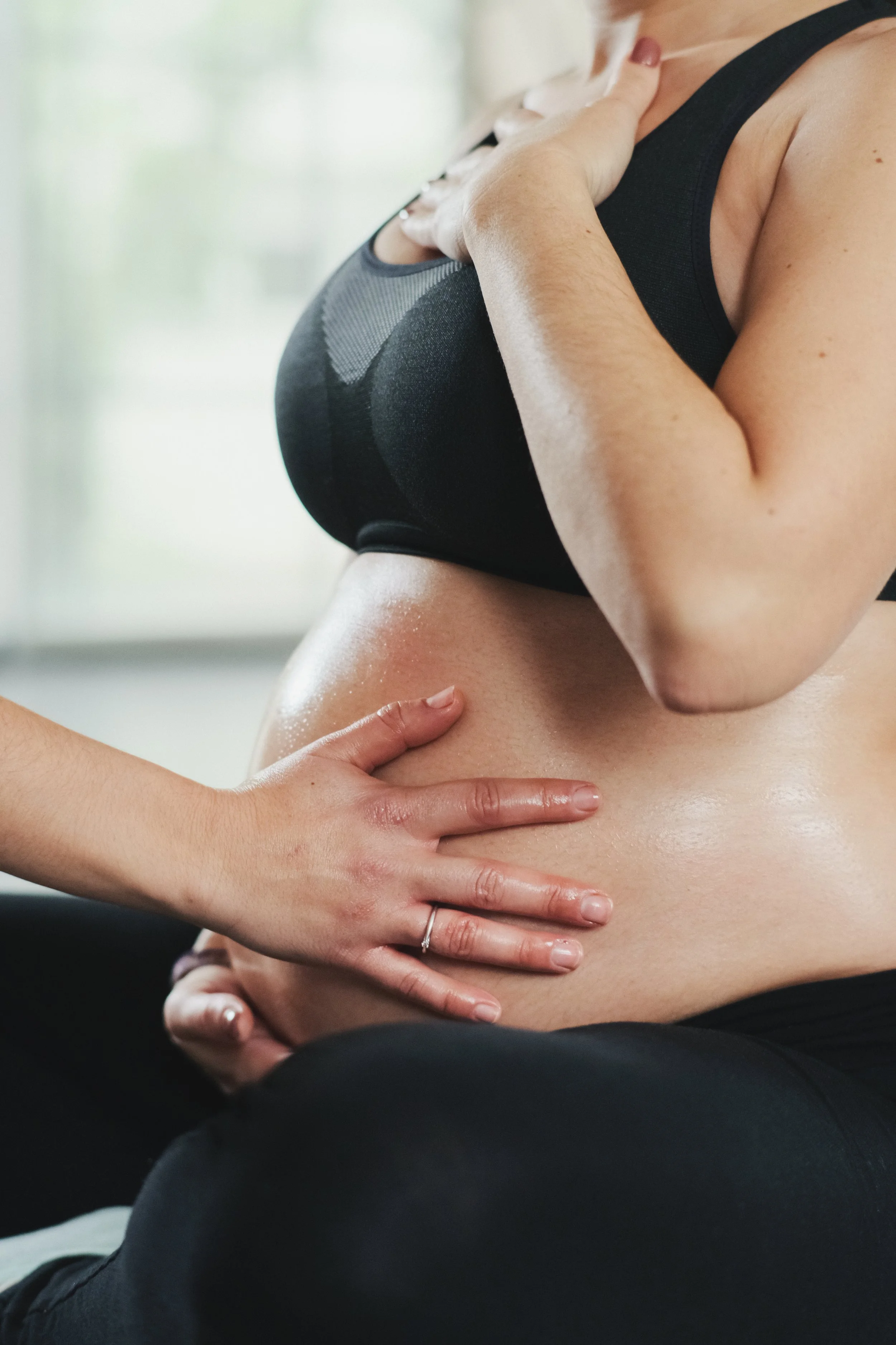 Una mujer con cabello rubio en ropa deportiva negra, sostiene su pecho en un lugar claro y brillante. Otra mano, de piel más clara y sin adornos, toca en su abdomen.