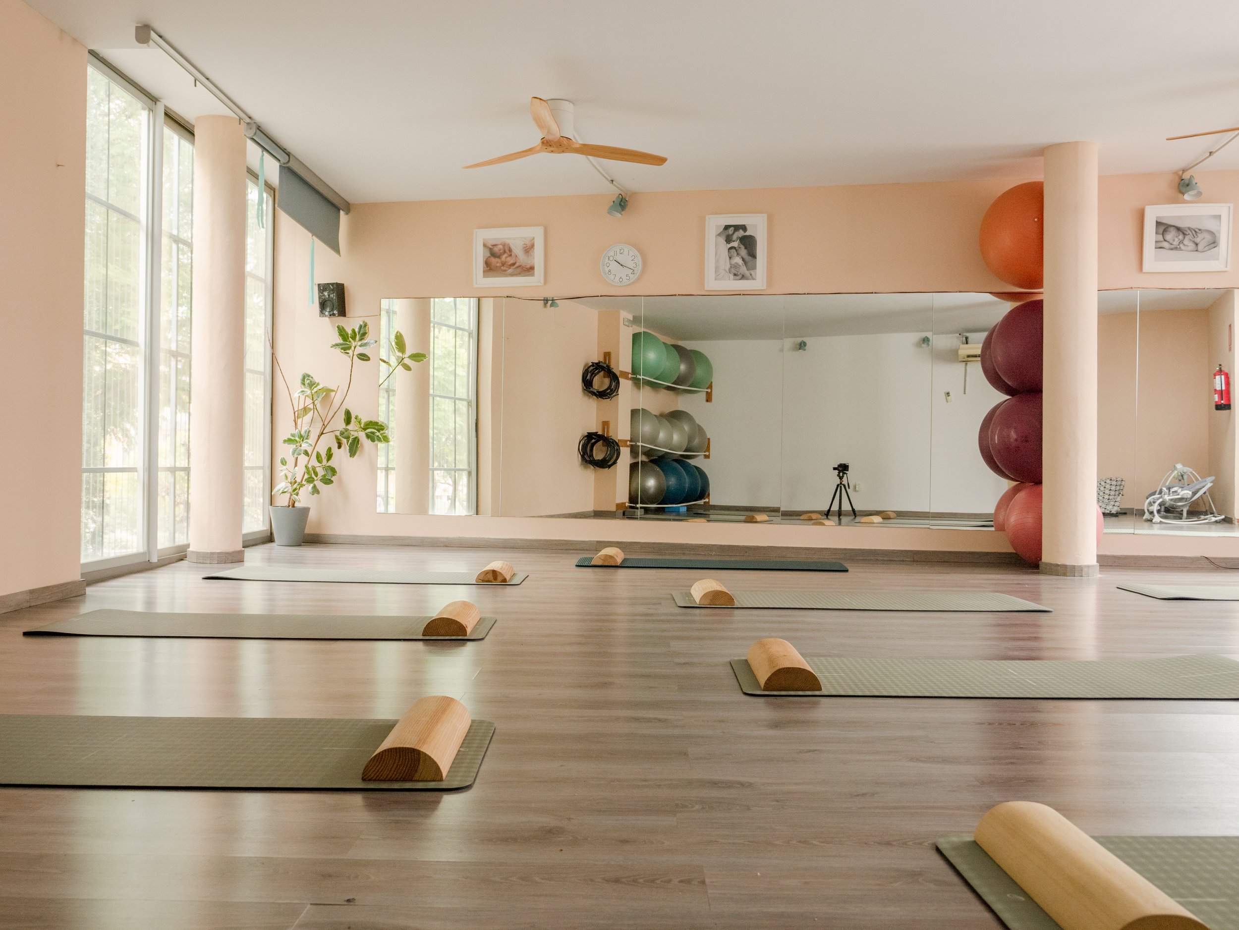 Sala de yoga con colchonetas y bloques de madera, paredes con espejos, pelotas de ejercicio, y ventanas grandes con cortinas blancas.