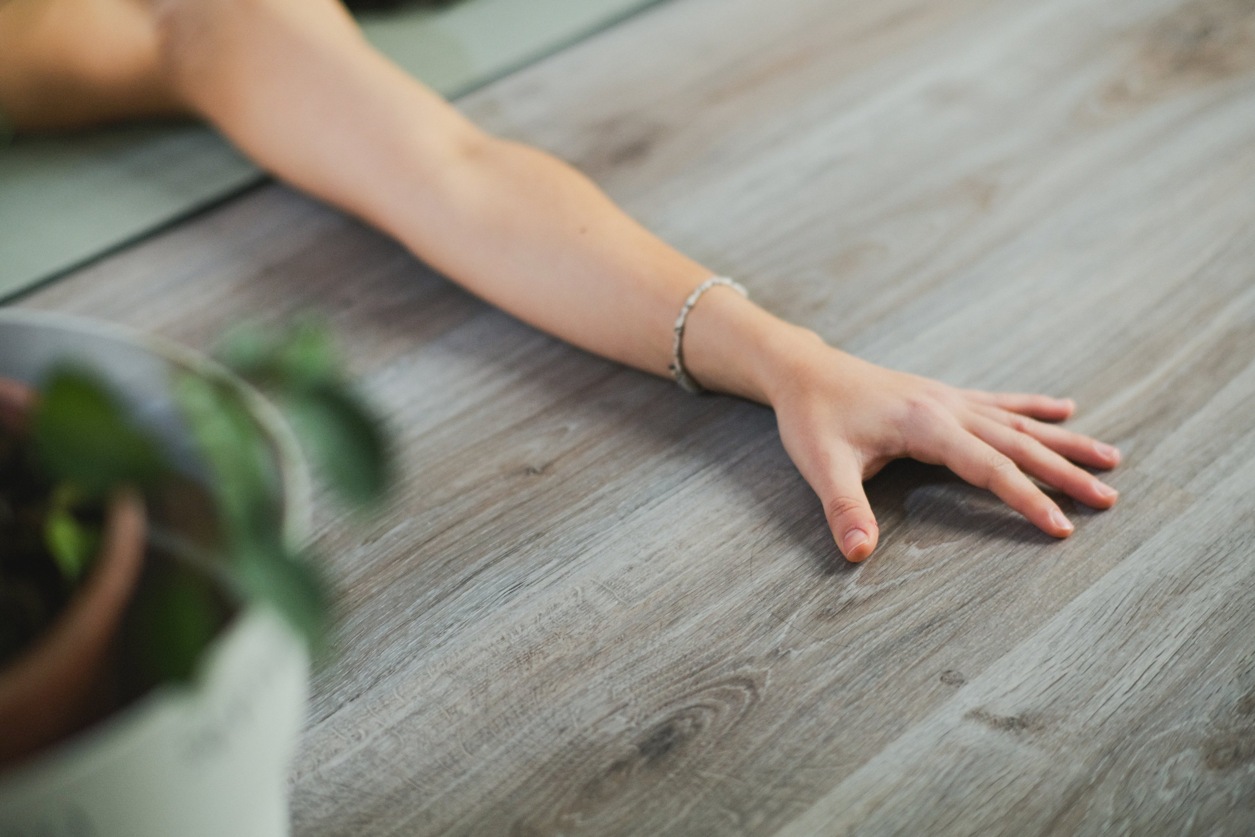 Persona en posición de estiramiento en el suelo, vista parcialmente de un brazo extendido con muñequera, en una superficie de madera, cerca de una planta en una maceta.