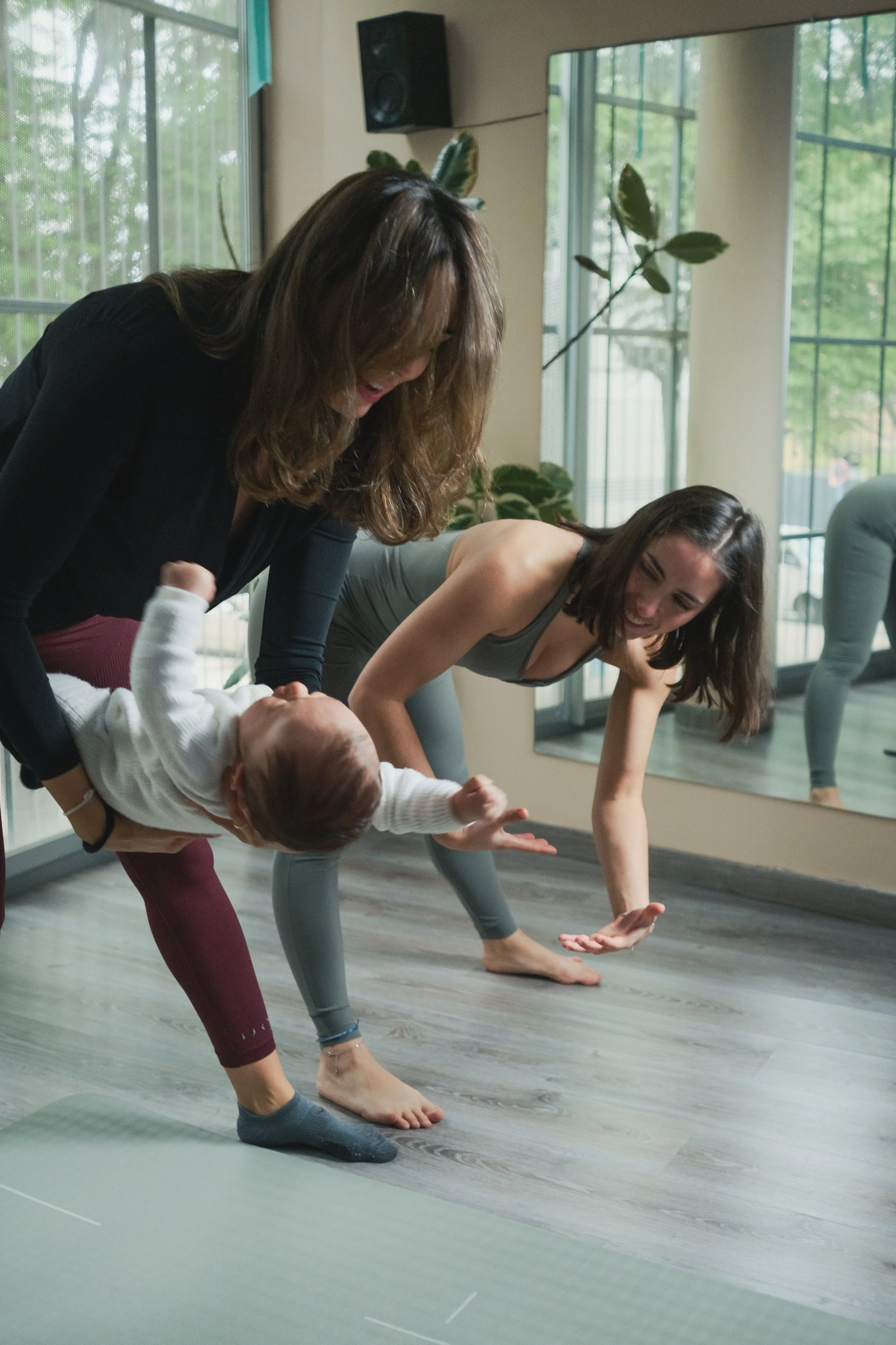 Dos mujeres y un bebé haciendo yoga en un estudio con paredes de vidrio y plantas en el fondo.