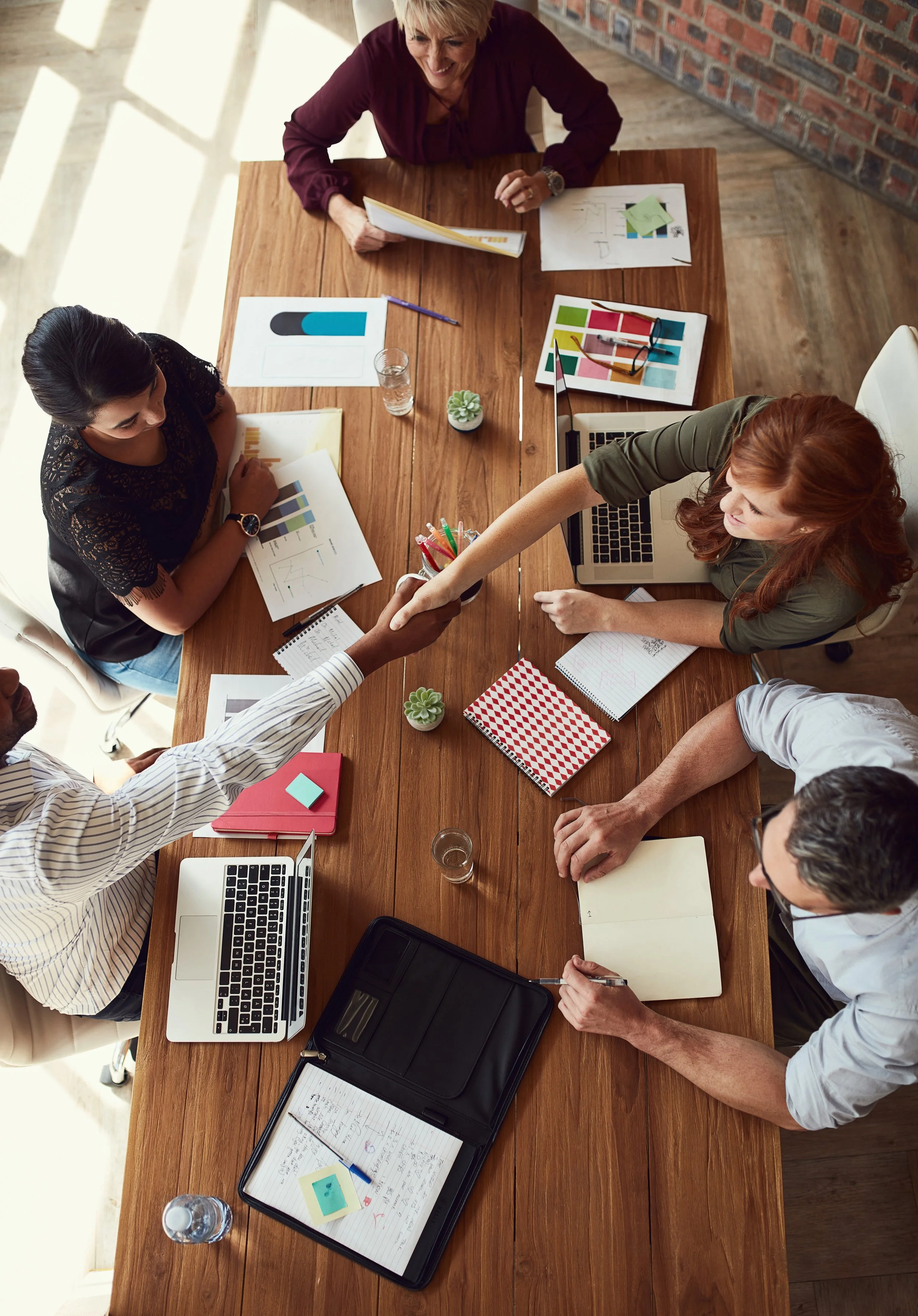 A group of six diverse professionals gathered around a wooden conference table, engaged in a collaborative meeting. The table holds laptops, documents with charts and graphs, notebooks, pens, sticky notes, and a small potted plant. A woman with red hair and a green shirt shakes hands with a man in a striped shirt, while others examine printed charts and take notes. Natural light from windows illuminates the scene.