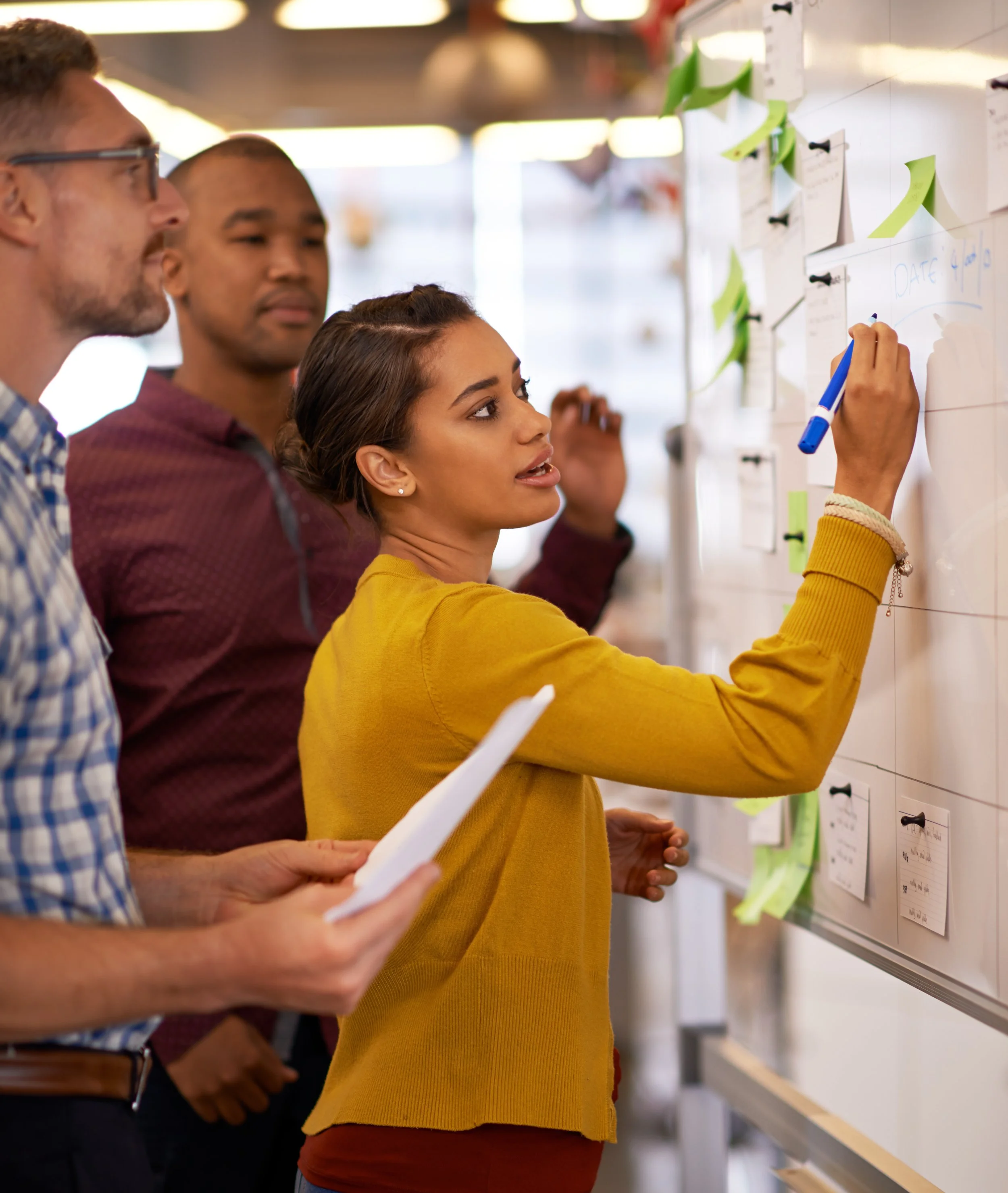 A group of people standing in front of a whiteboard, with a woman in a yellow sweater writing notes on the board using a blue marker. They are in a room with lockers and notes pinned to the wall.