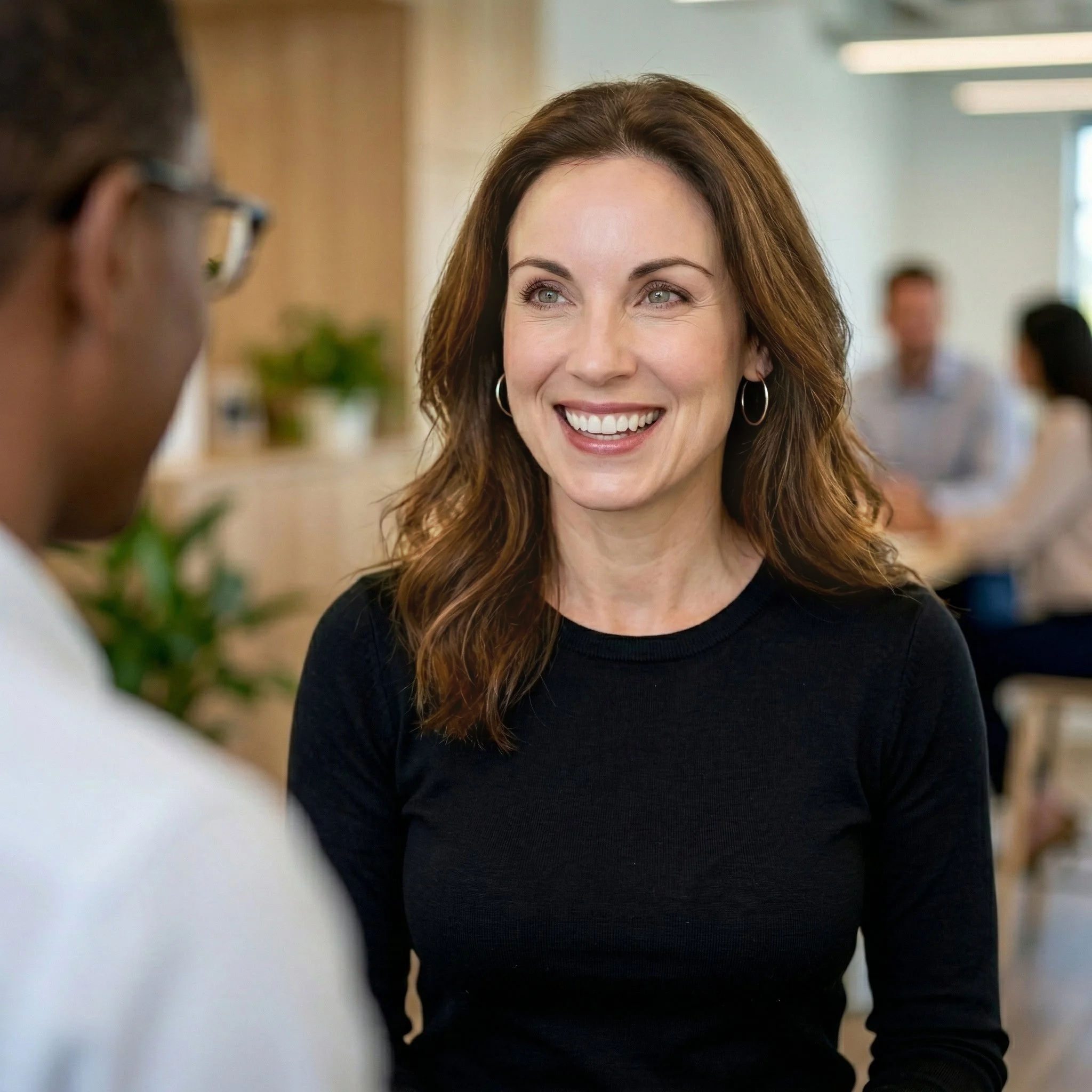 Smiling woman with brown hair and hoop earrings talking to a man wearing glasses in a bright indoor setting.