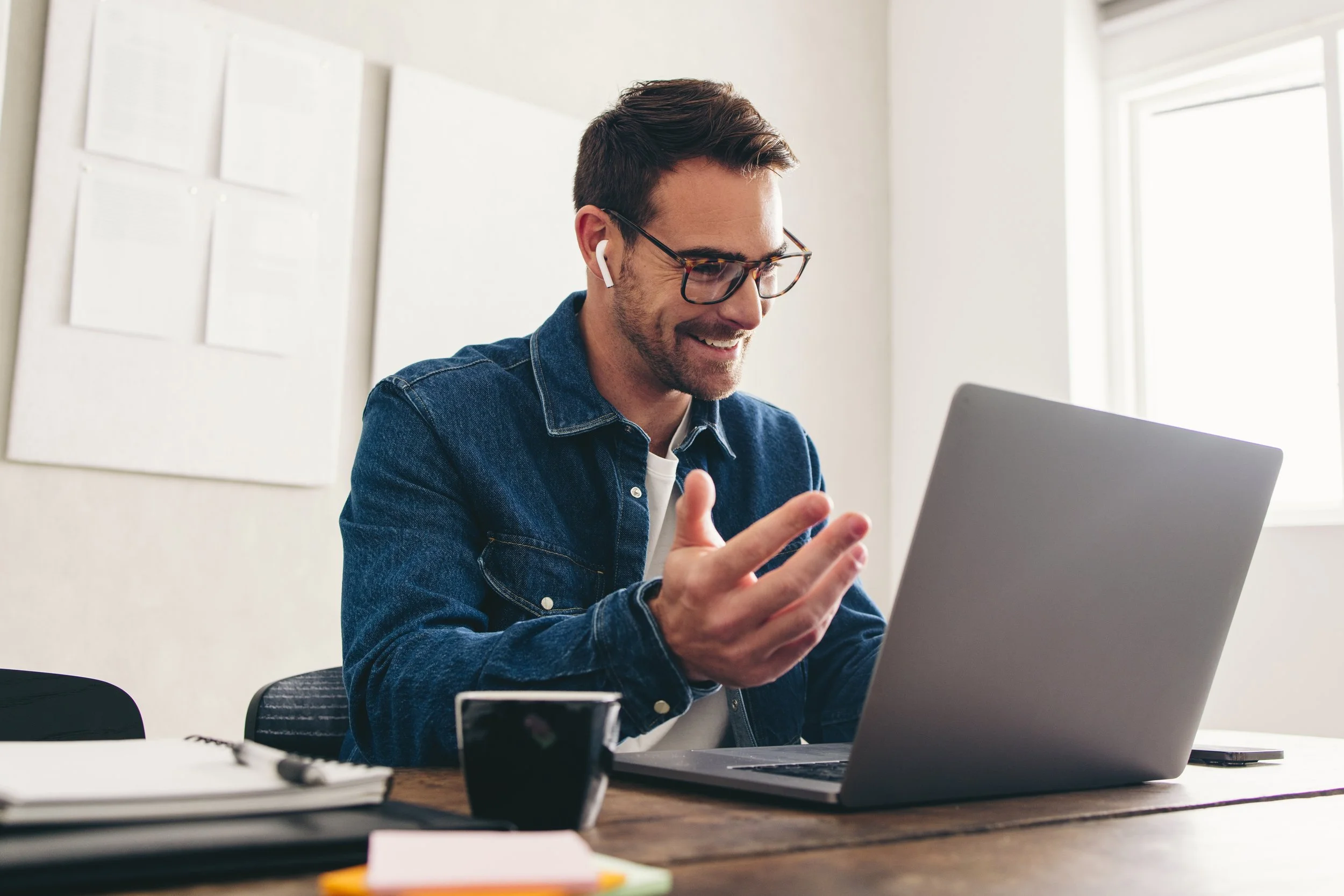 A man with dark hair, glasses, and a beard is smiling and talking on his smartphone while sitting at a desk with a laptop, paper, and a pen in a bright office space.