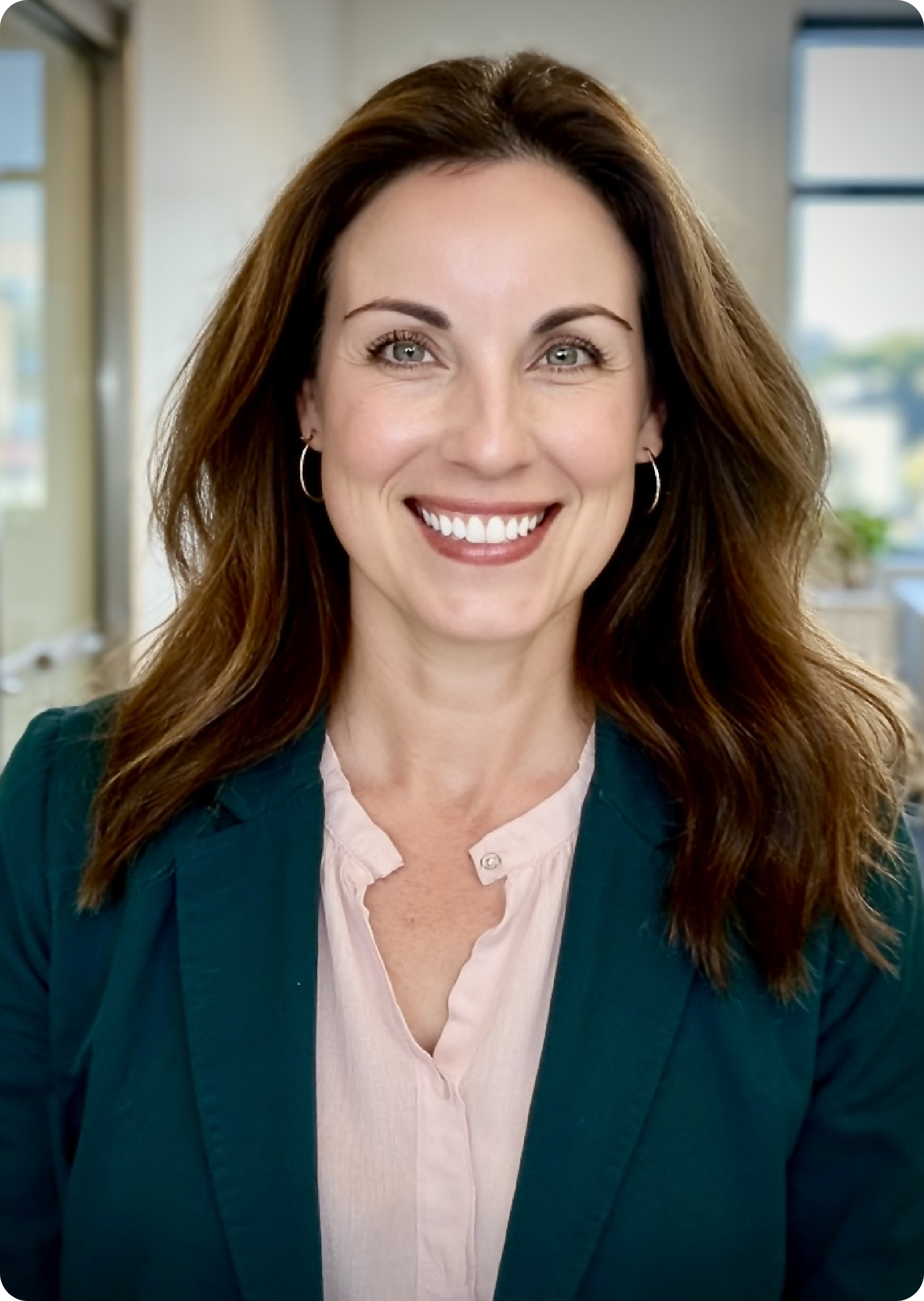 Close-up of a woman with brown hair, green eyes, and hoop earrings, smiling in an office with windows in the background.