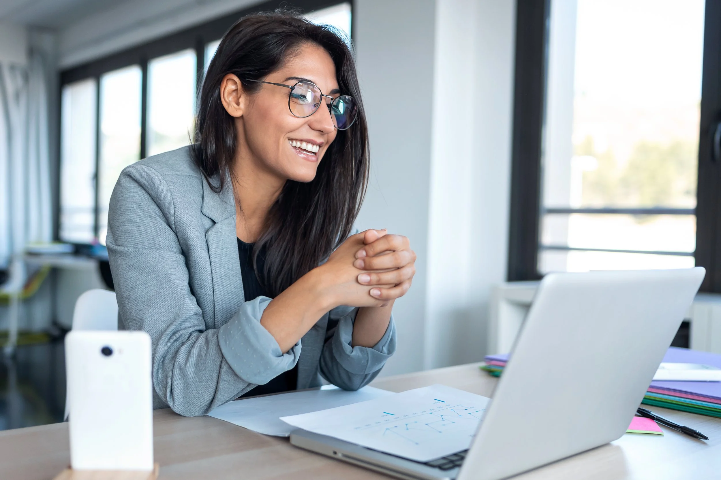 A woman with dark hair and glasses, smiling while sitting at a desk with papers, a laptop, and colorful sticky notes in a bright office.