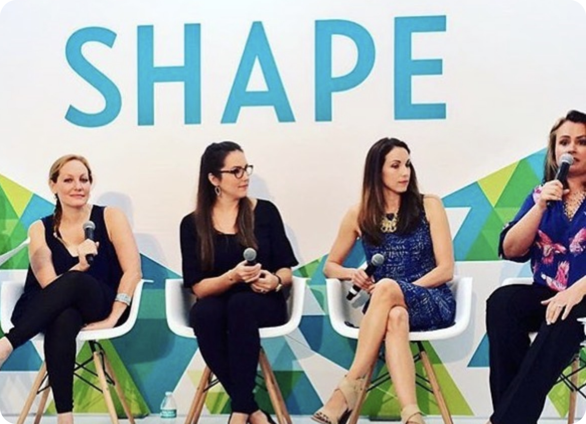 A group of women sitting on chairs with a large sign behind them that reads 'SHAPE' in blue letters.