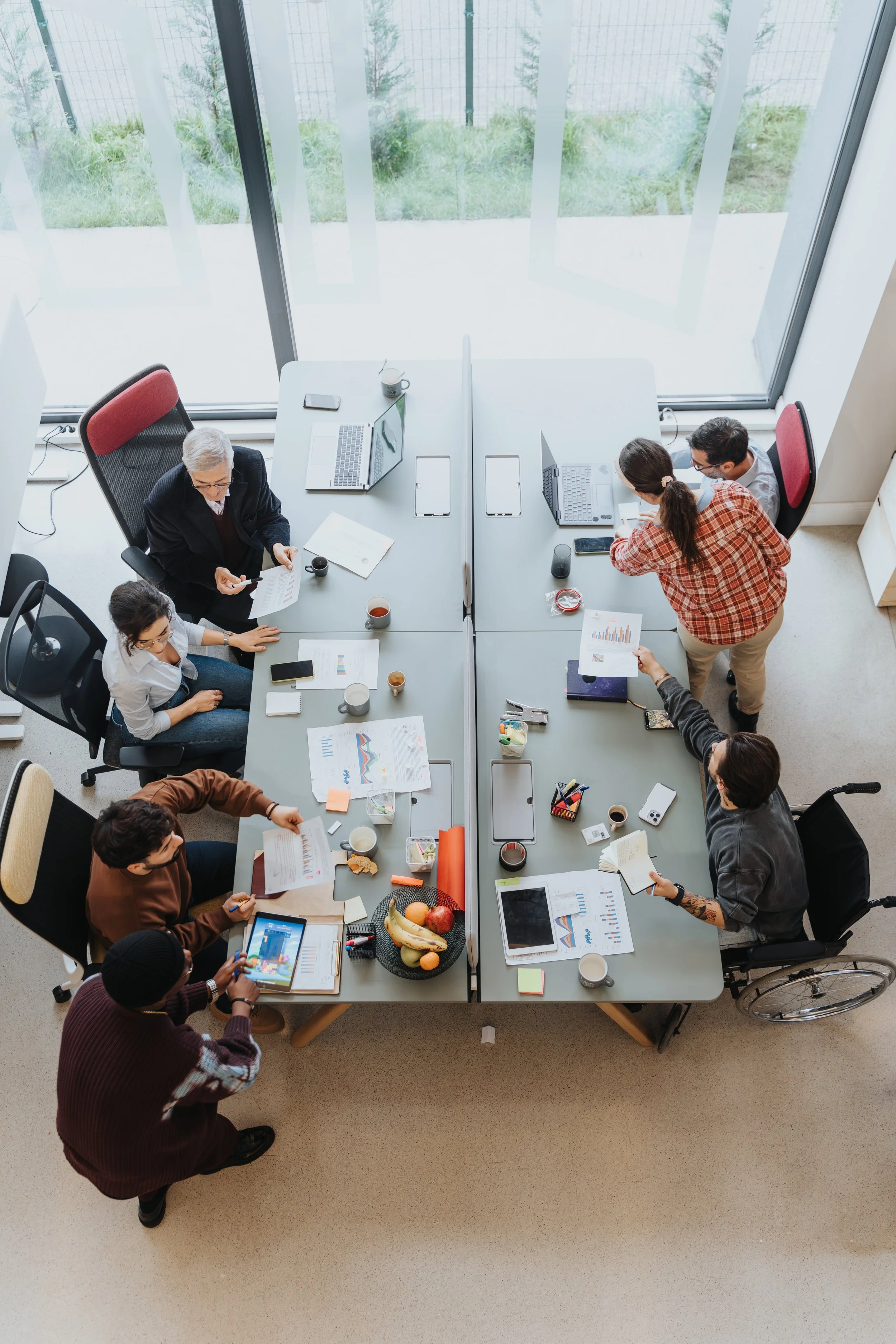 An overhead view of a diverse group of people working together in a modern office conference room with large windows, laptops, documents, and various snacks and drinks on the table.