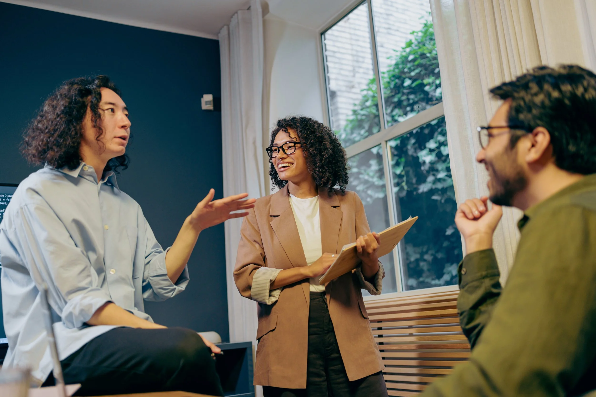 Three people having a discussion in a bright room with large windows and beige curtains. One woman with curly hair and glasses is smiling and holding a clipboard, another woman with curly hair is speaking, and a man with glasses and dark hair is listening.
