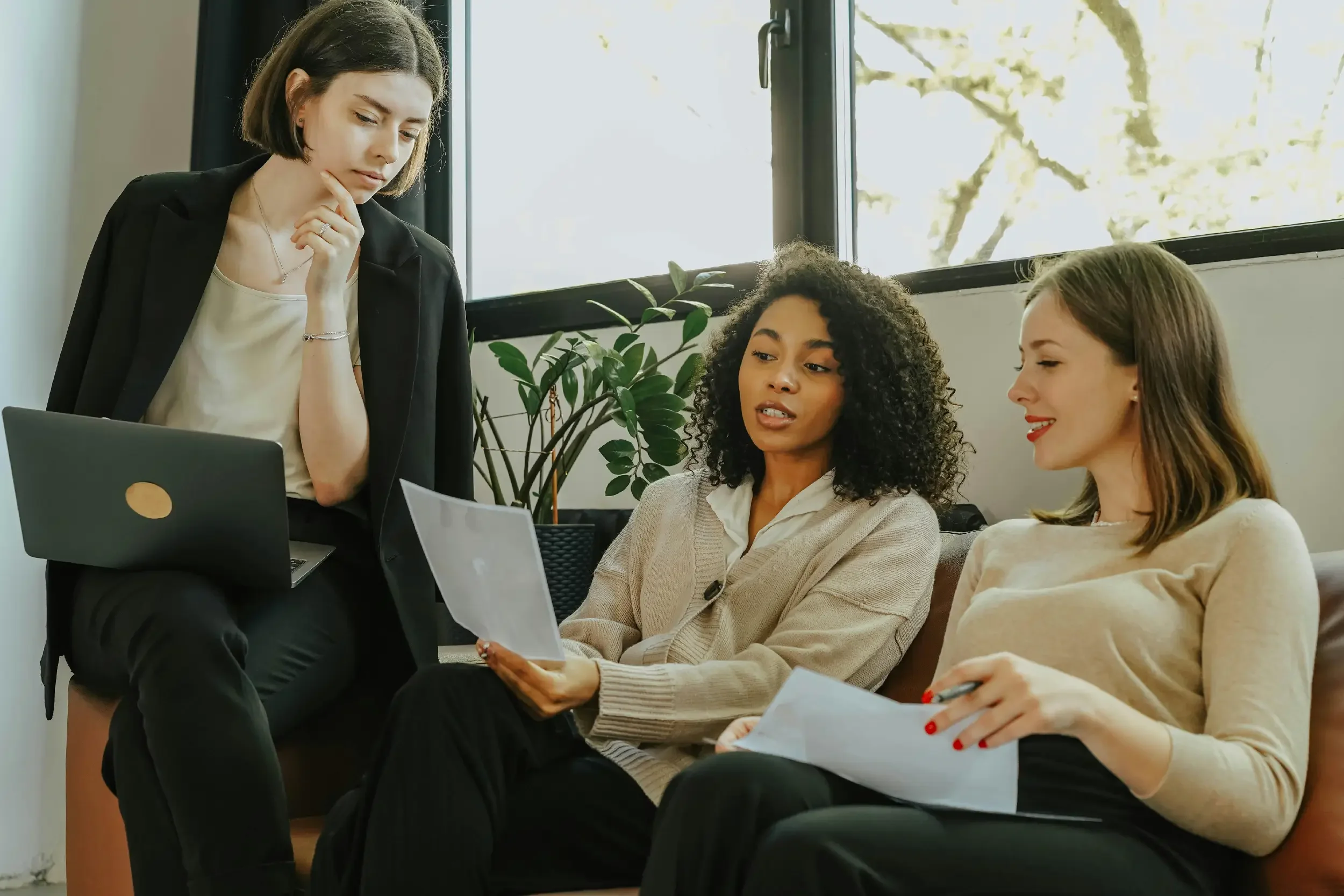 Four women in a office or meeting room. One woman is standing and looking at the laptop while the other three are seated, holding papers and looking at the standing woman. Large windows with trees outside are visible in the background.