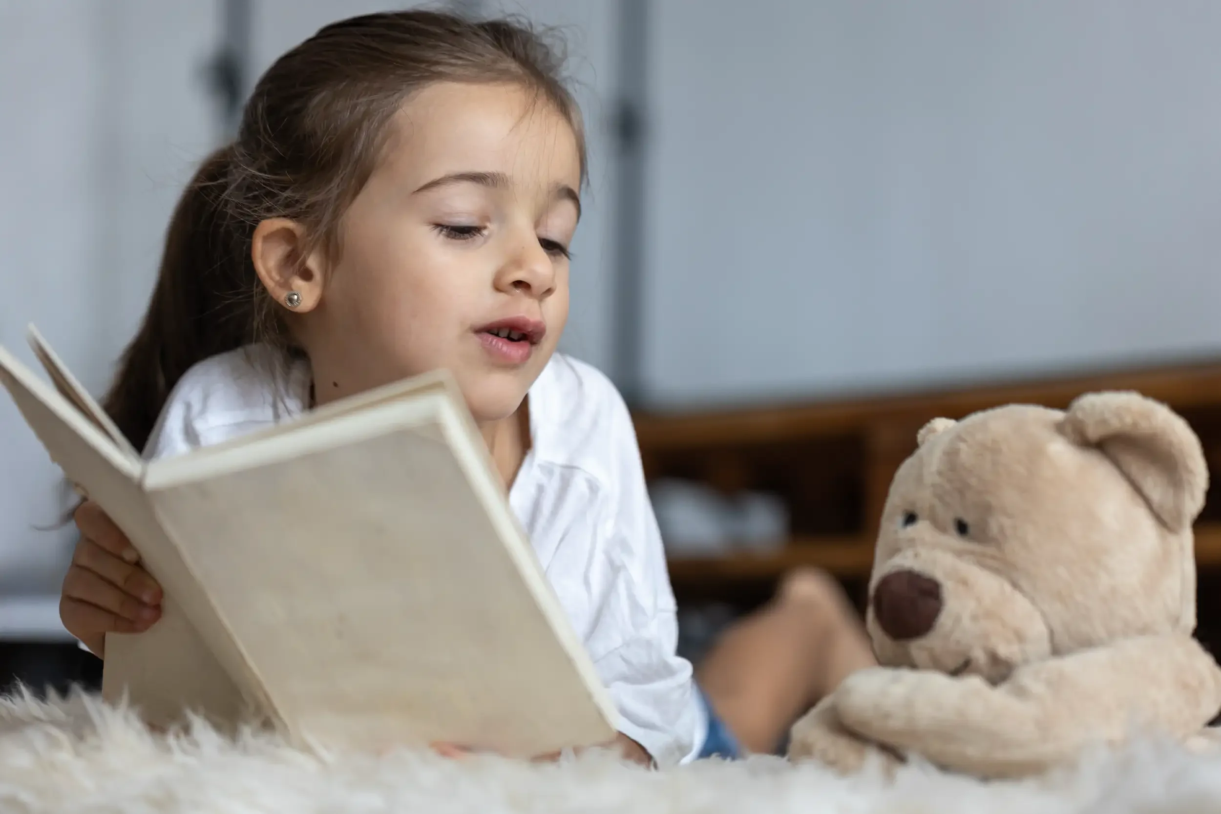 A young girl with brown hair and earrings reading a book while sitting on a white fluffy surface, with stuffed teddy bears nearby.