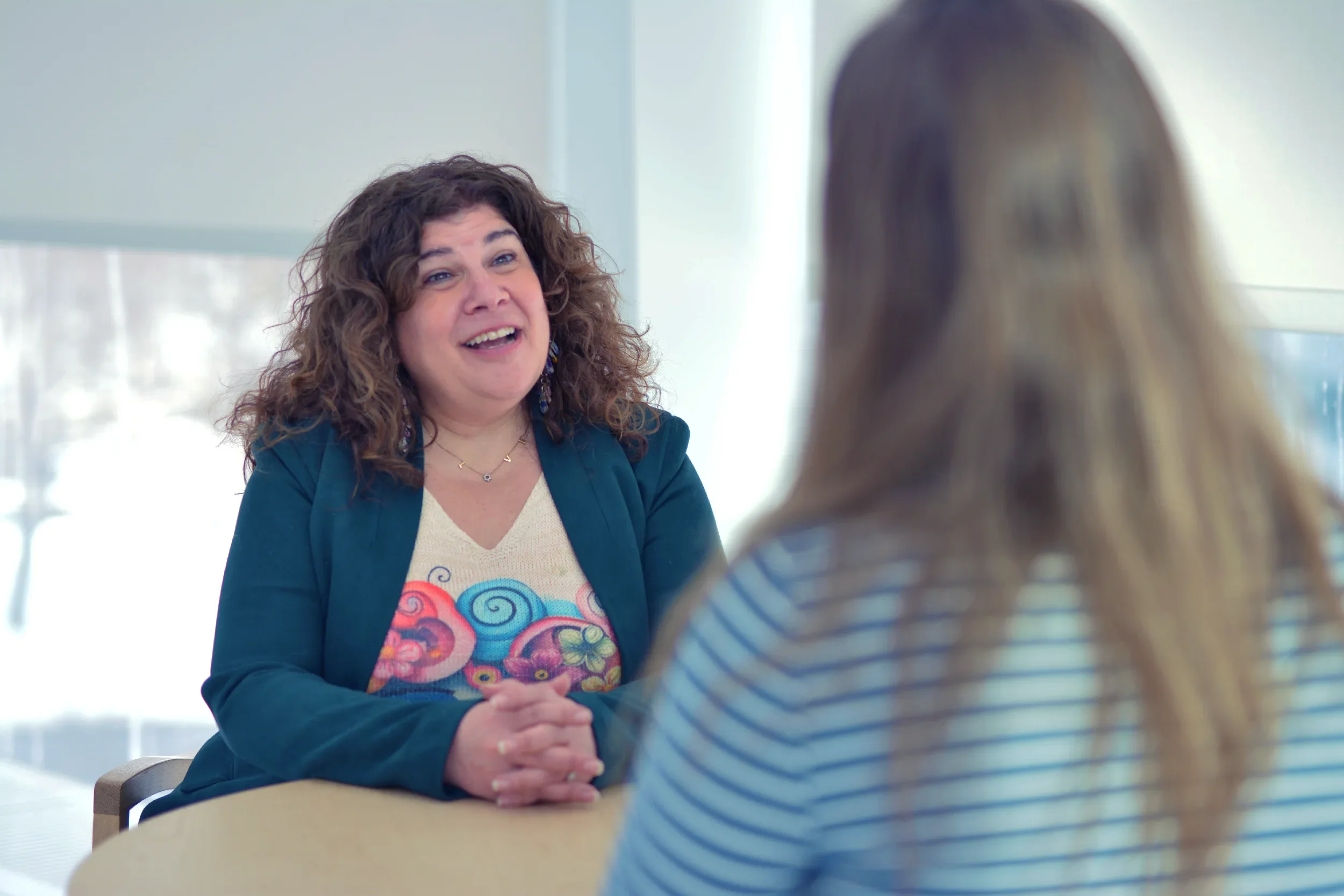 A woman with curly brown hair, wearing a colorful RAINBOW LIVIN graphic T-shirt and a dark blazer, smiling and holding hands with a person in a striped shirt in a bright, well-lit room.