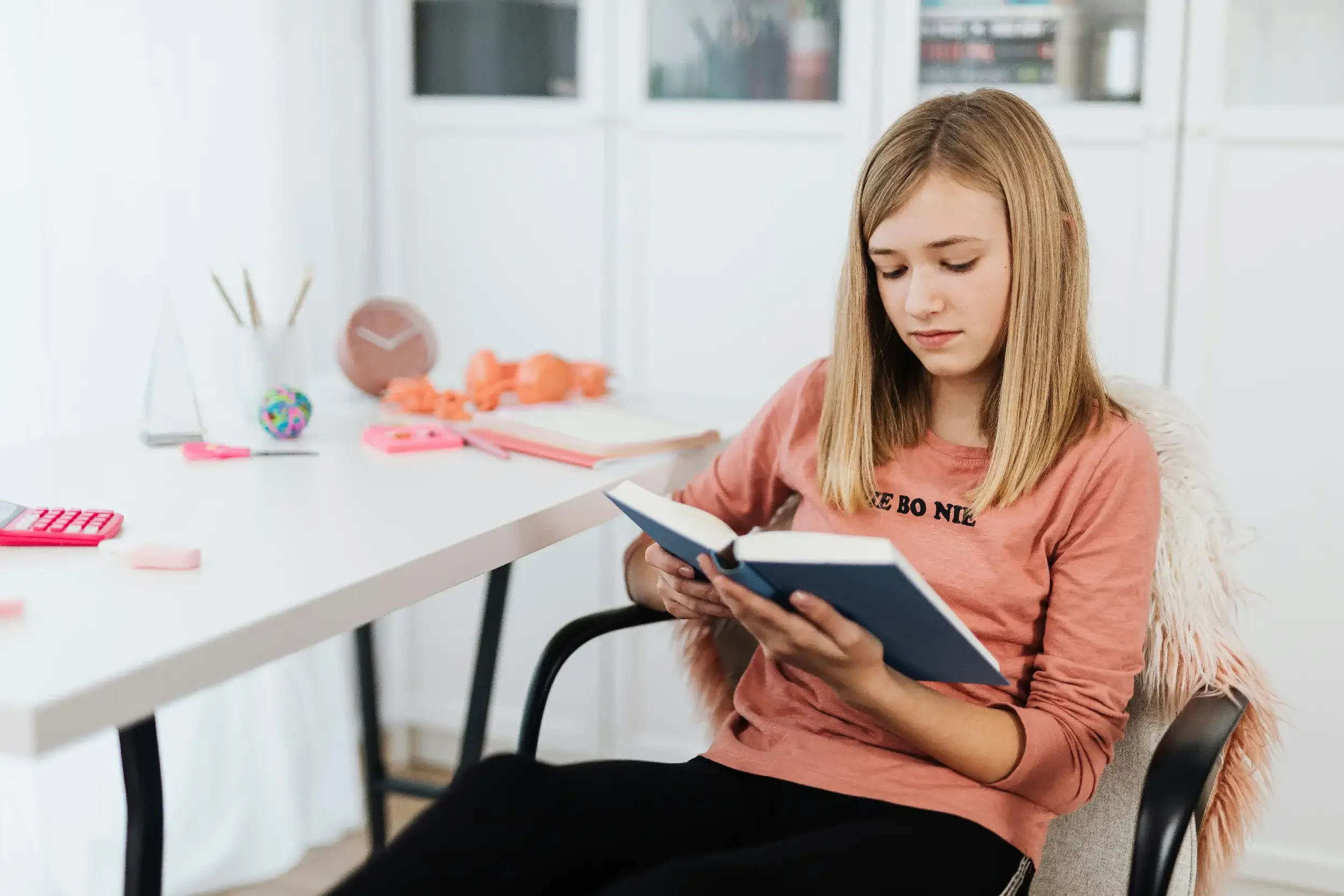 A young girl with blonde hair and a pink shirt sitting on a chair at a white desk reading a book.