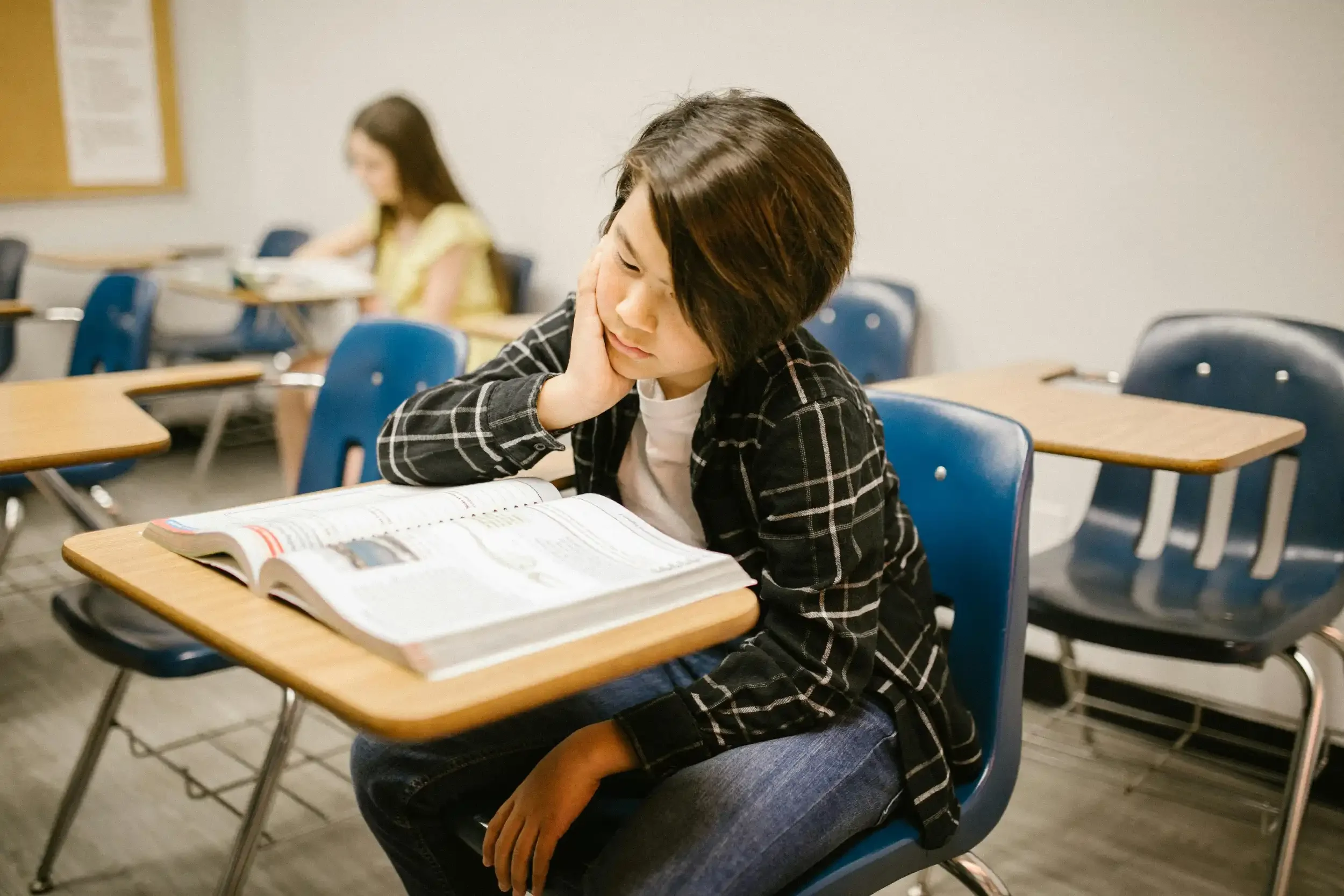 A young woman sitting at a desk, resting her head on her hand, appearing to be tired or bored, with an open textbook in front of her in a classroom. A second person is in the background, blurred, also seated at a desk.