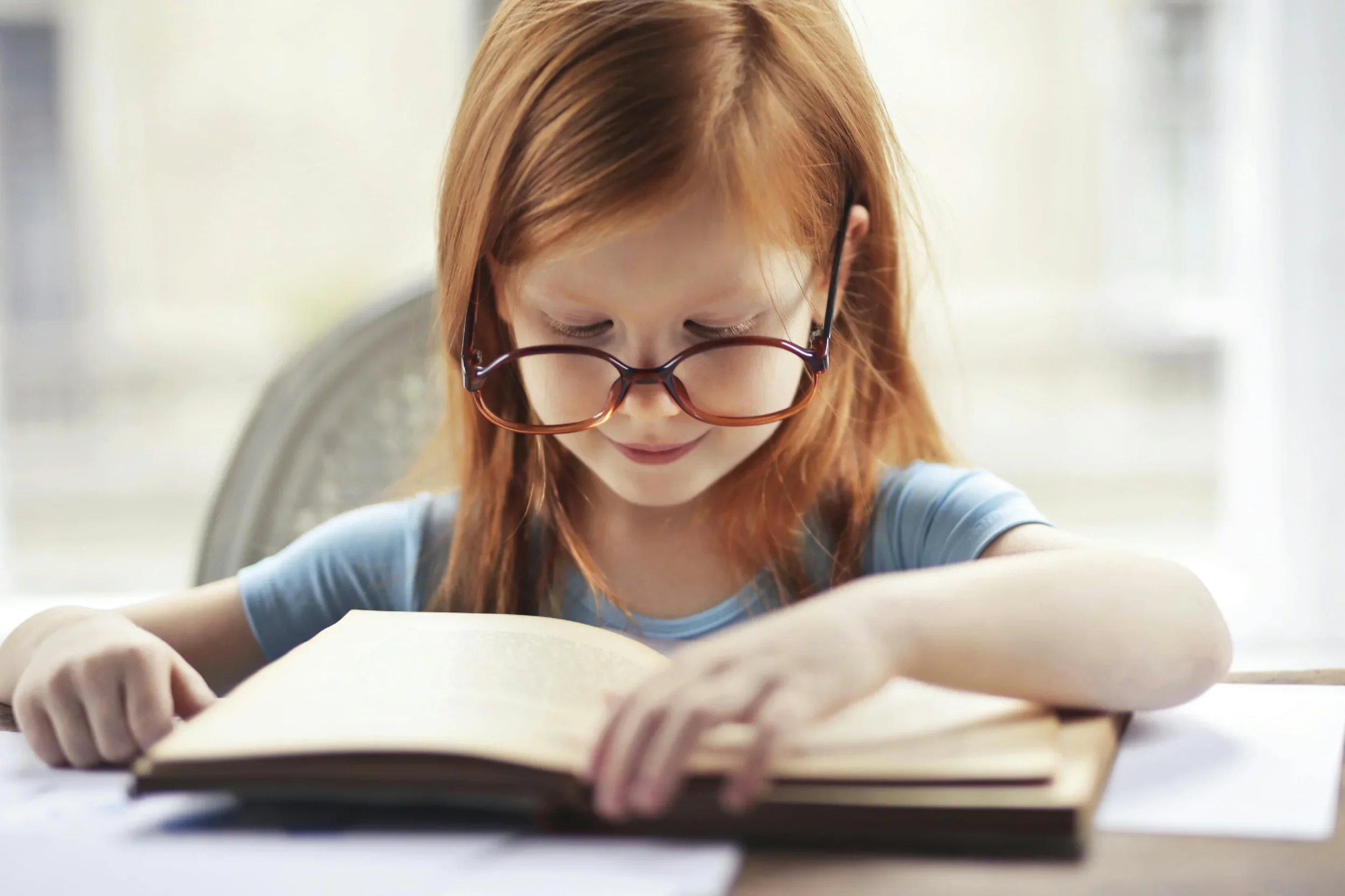 A young girl with long red hair wearing glasses reading a book at a table.