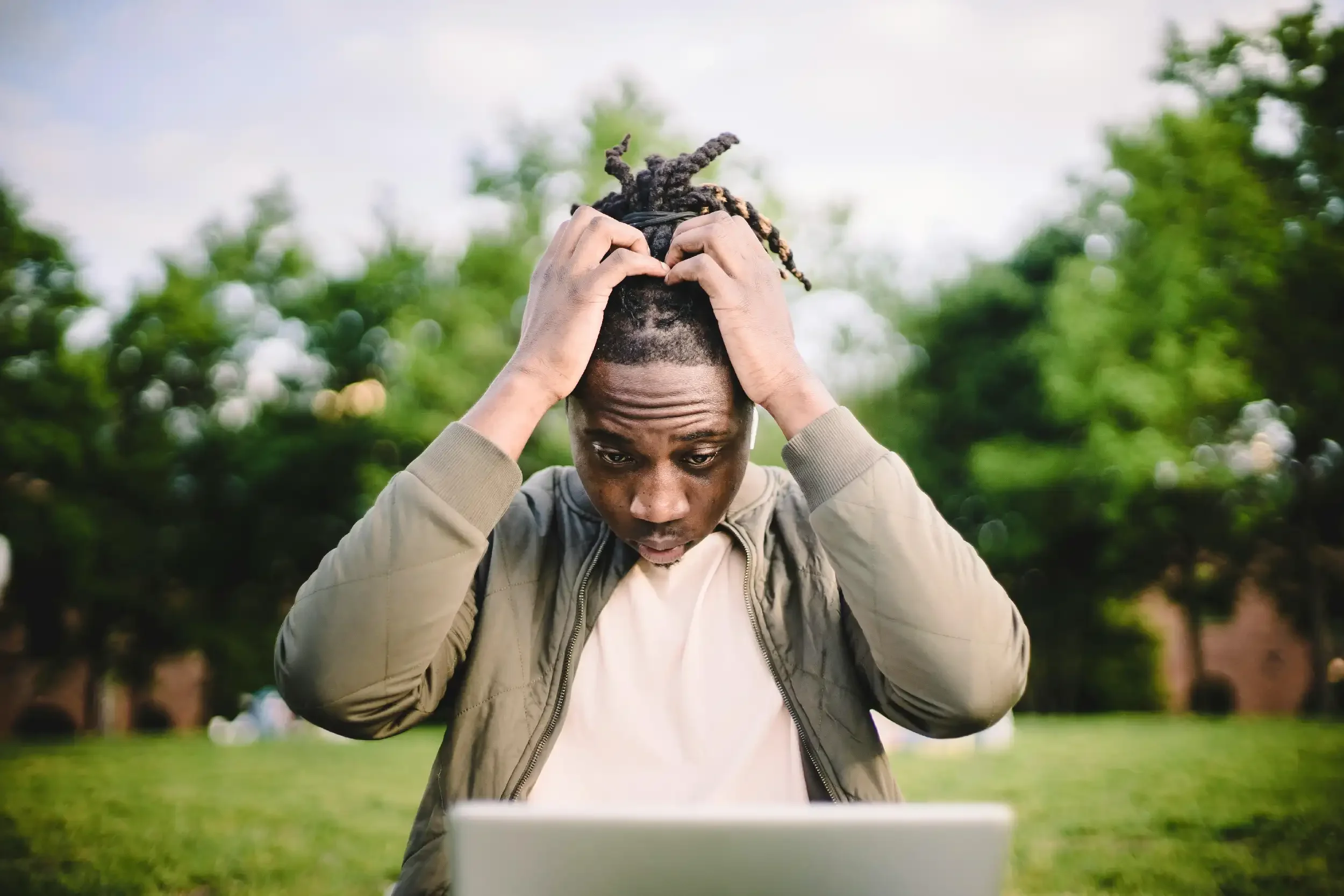 A young man with dreadlocks adjusts his hair while sitting in a park, looking down at a laptop with green trees and grass in the background.
