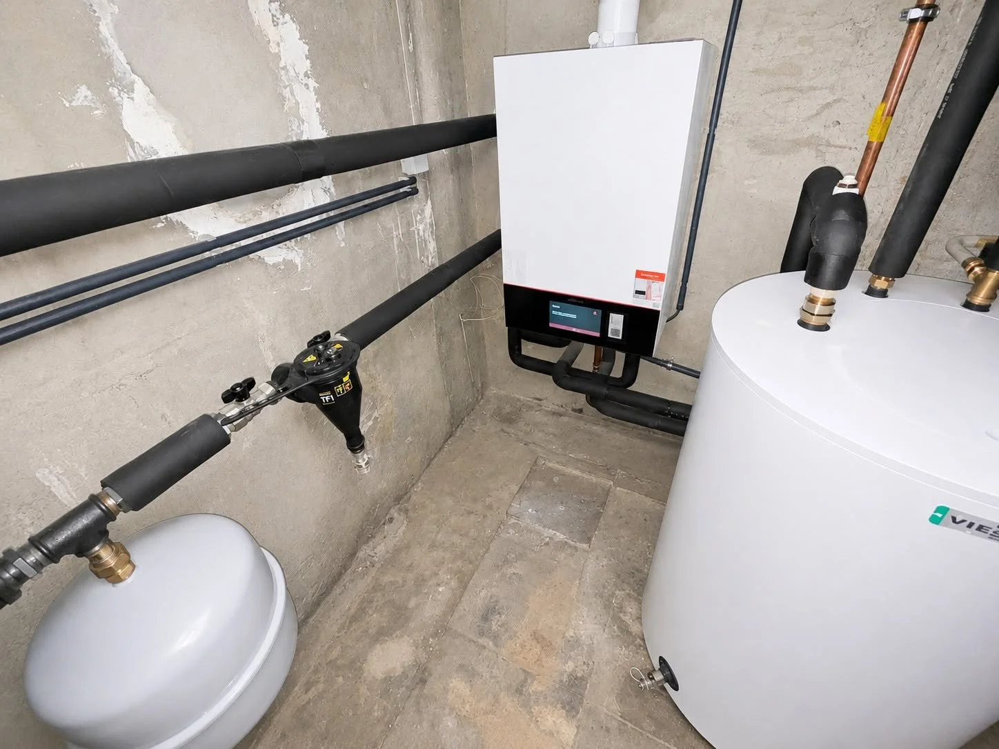 Salle technique avec une chaudière, des tuyaux en cuivre et en PVC, un panneau de contrôle numérique, et un ballon d'eau chaude