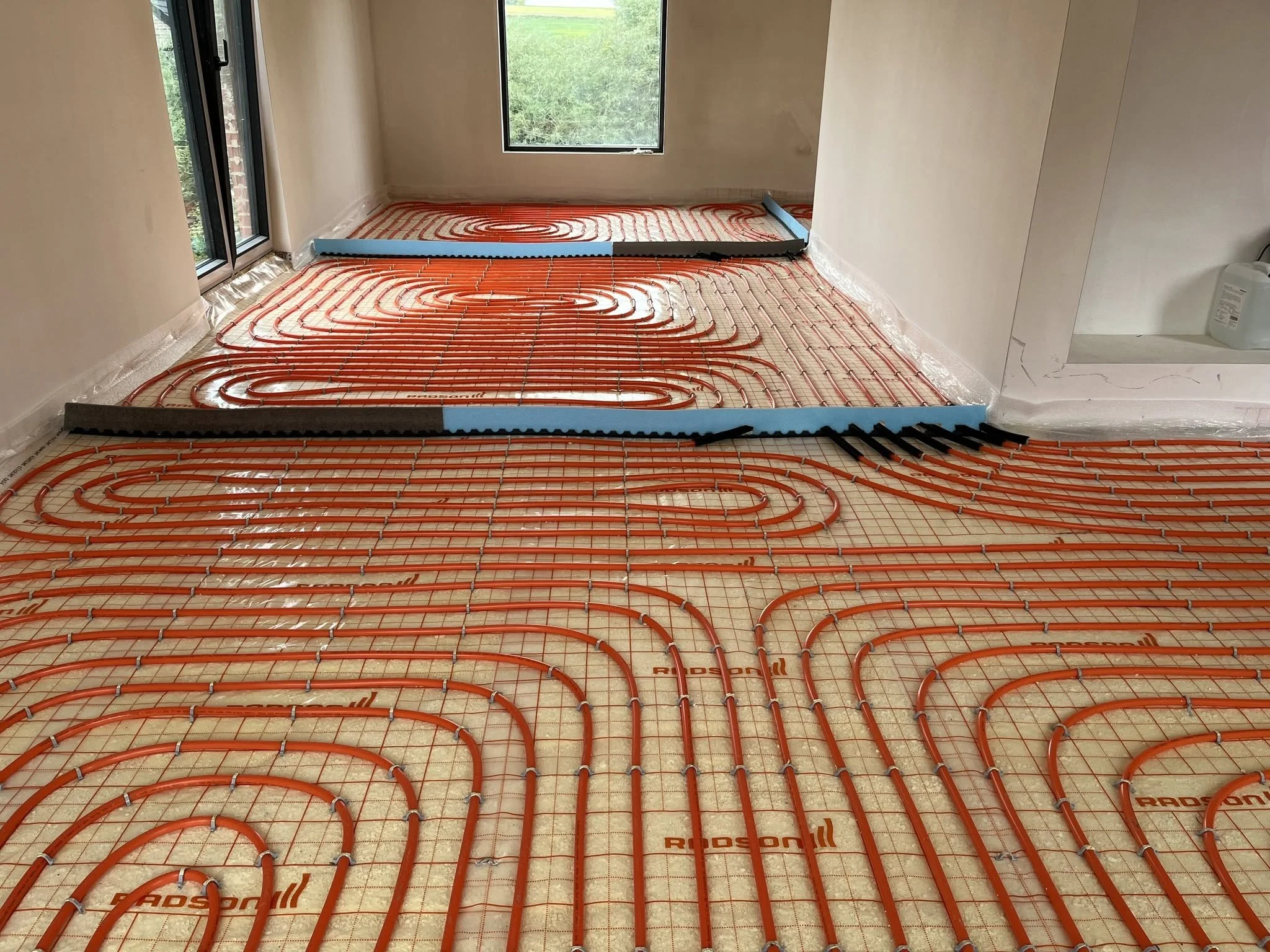 Installation de chauffage par le sol avec des tuyaux orange disposés en spirale, aspect en cours d'installation dans une pièce avec fenêtres et murs clairs.