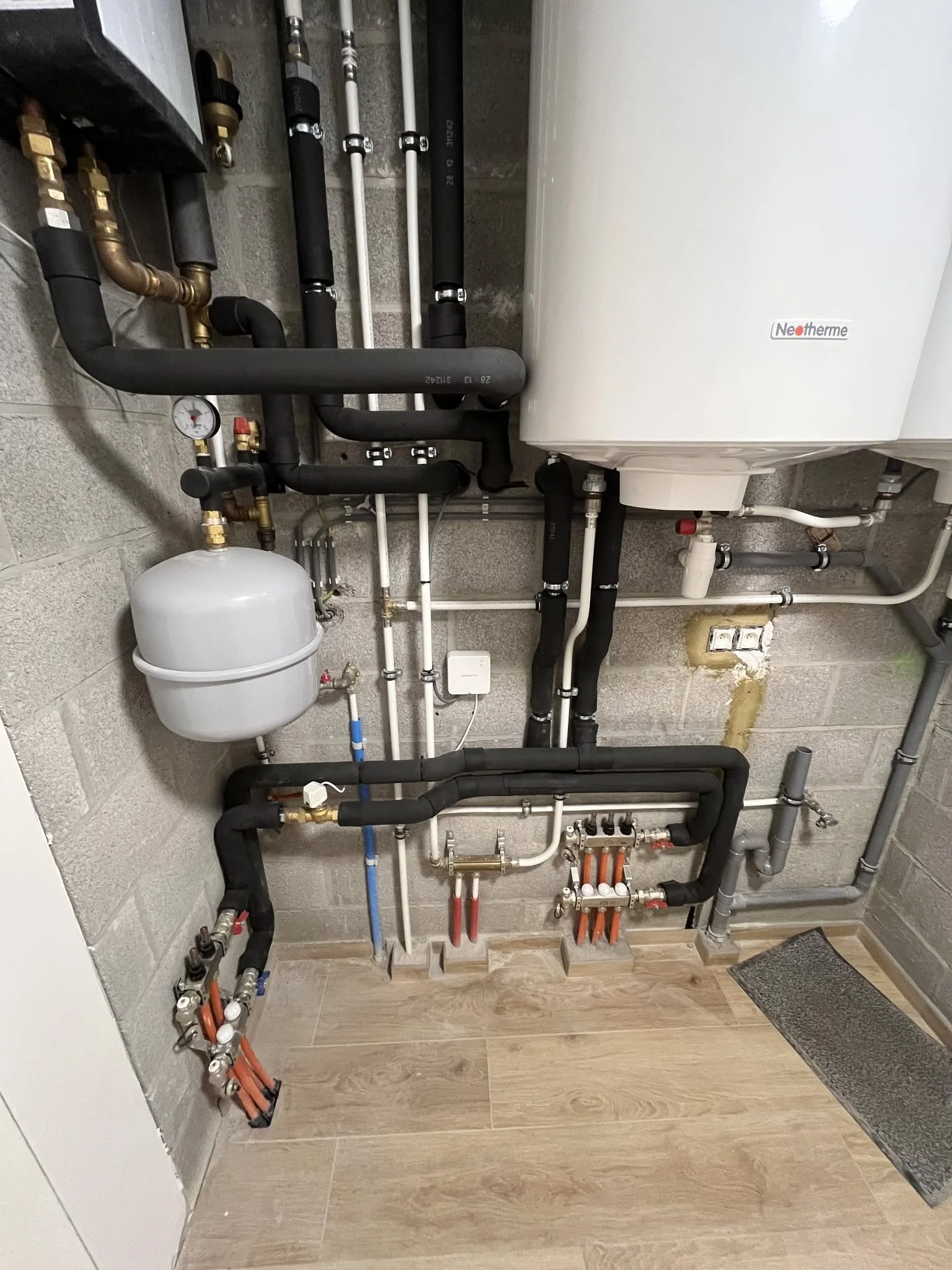 Salle technique avec tuyaux de plomberie, un ballon d'eau chaude, un thermomètre et divers raccords, fixés sur un mur en béton et posé sur un sol en bois.