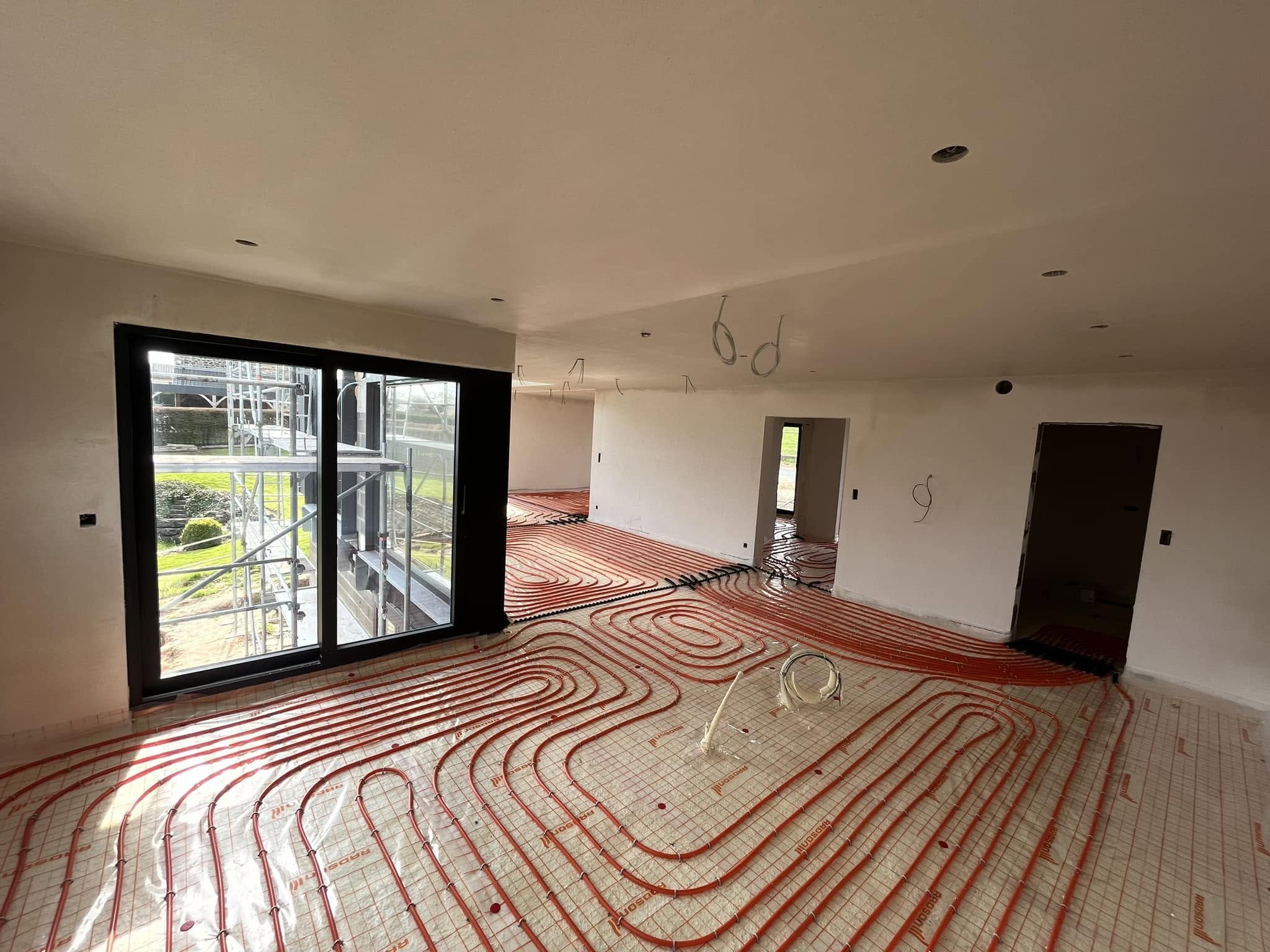Intérieur d'une maison en construction avec le sol radié par un système de chauffage par le sol, plomberie en cours d'installation, grande baie vitrée laissant entrer la lumière, murs en cours de peinture.