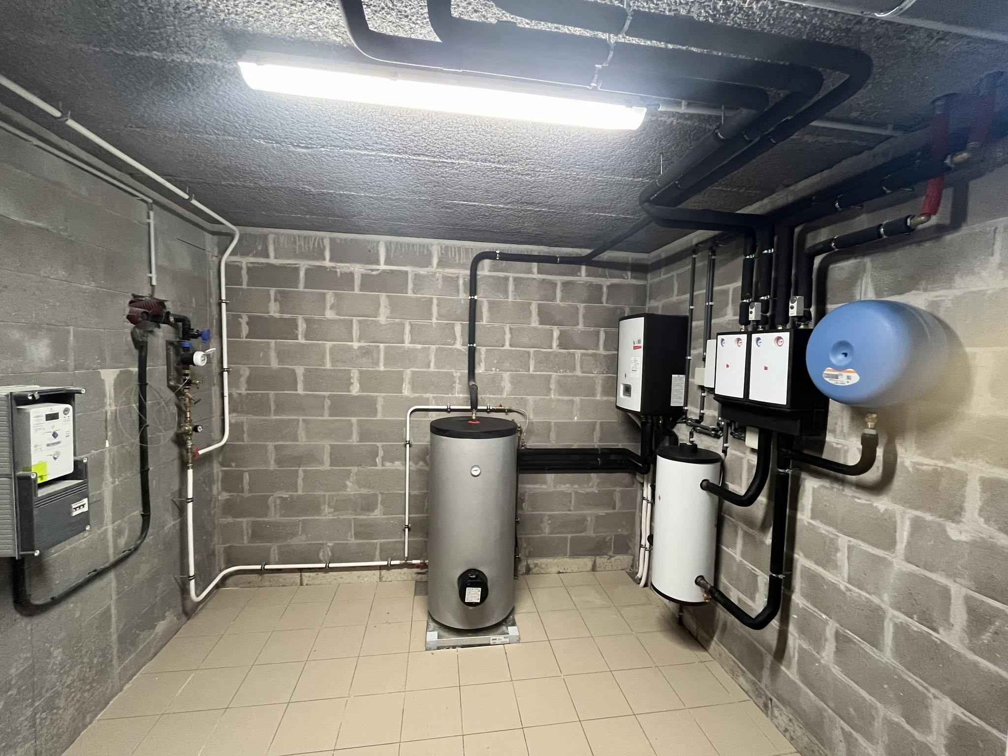 Salle technique avec équipements de plomberie, conduits et chaudières contre un mur en briques.