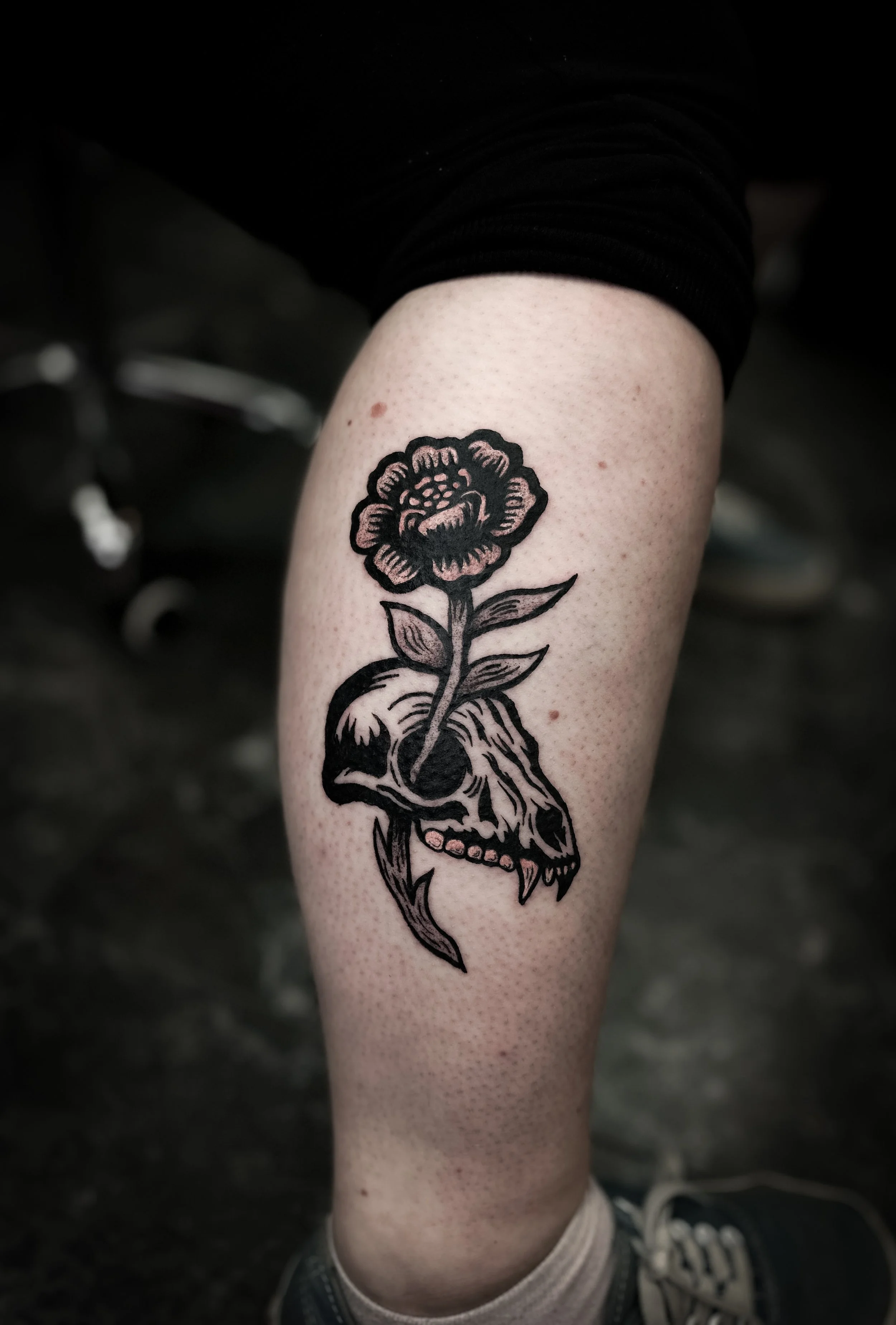 Tattoo of a skull with a flower growing out of it on a person's leg in an inkblock or woodcut style. 
