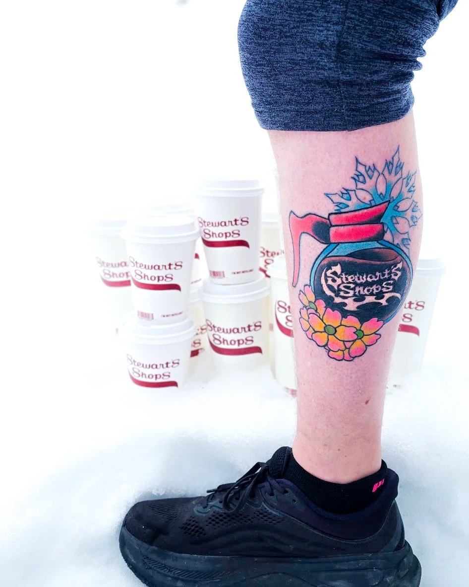 A person's leg with a tattoo of a heart-shaped explosive labeled 'Stewart's Shops', surrounded by flowers, over a layer of snow with Stewart's Shops cups in the background.